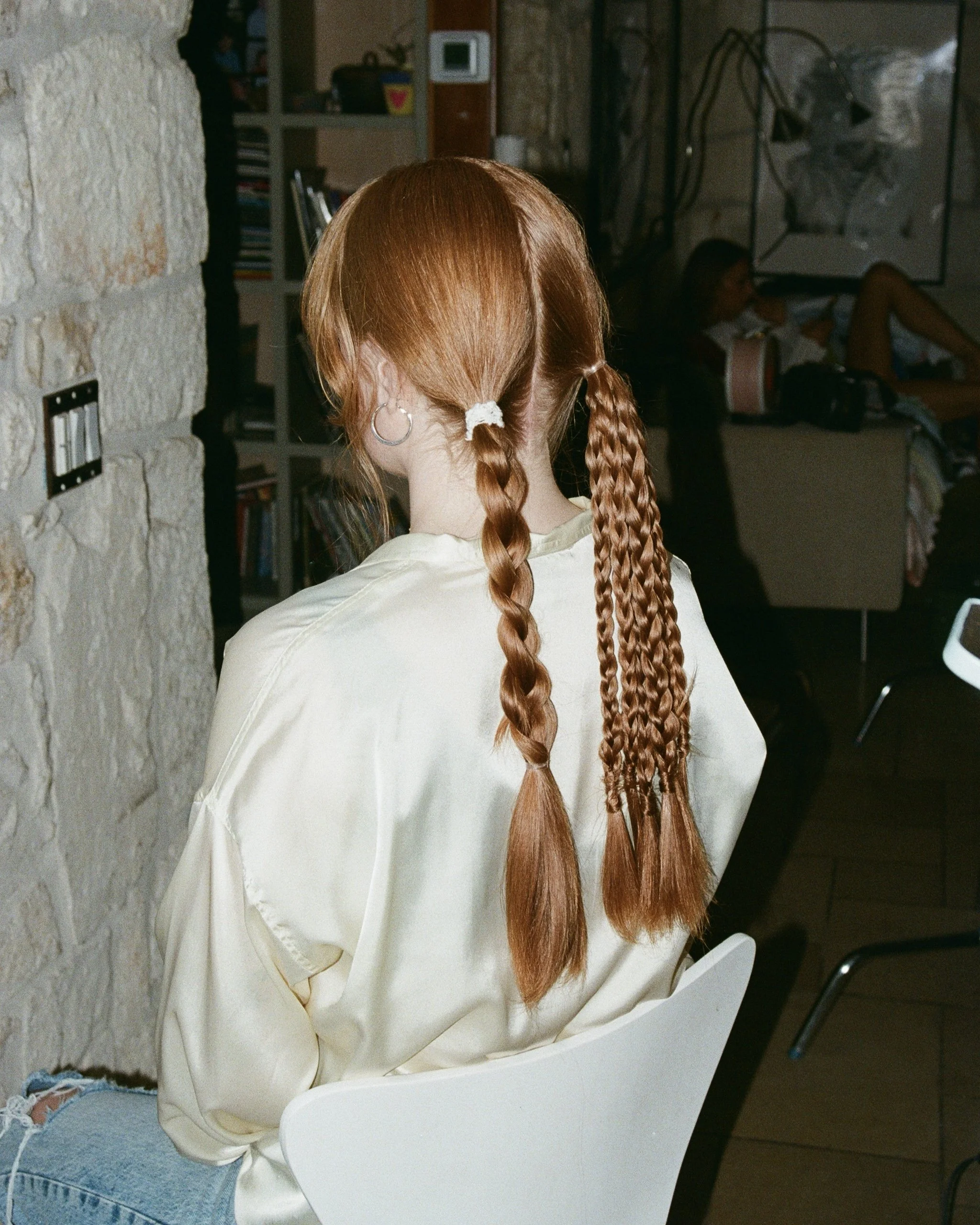 Back of a woman with red hair styled in two thick braids, wearing a cream satin blouse, sitting in a modern indoor setting with other people in the background.