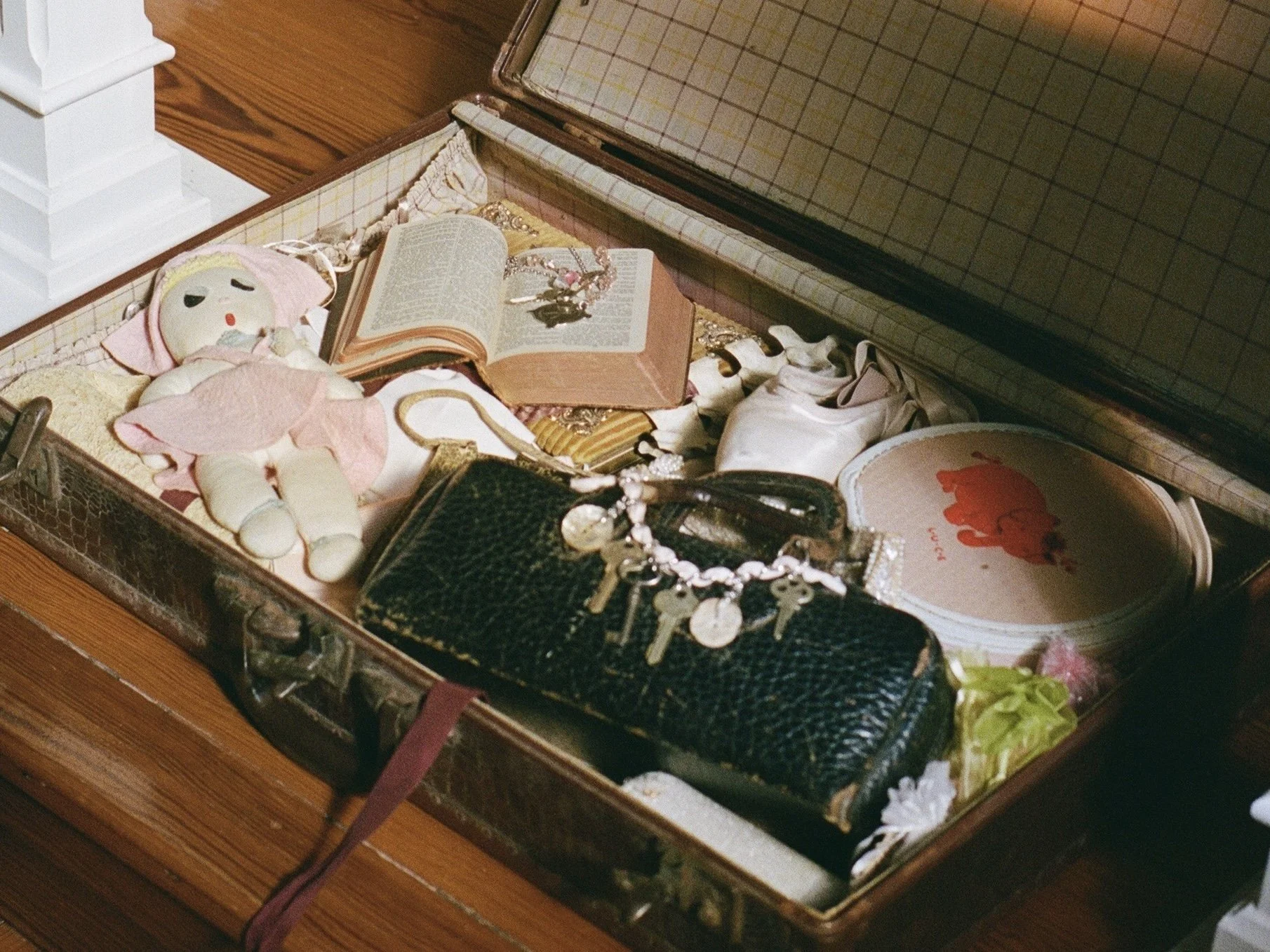 Open vintage jewelry box containing a stuffed doll, an open book, a black textured purse with jewelry, a round plate with a red design, and various small accessories.