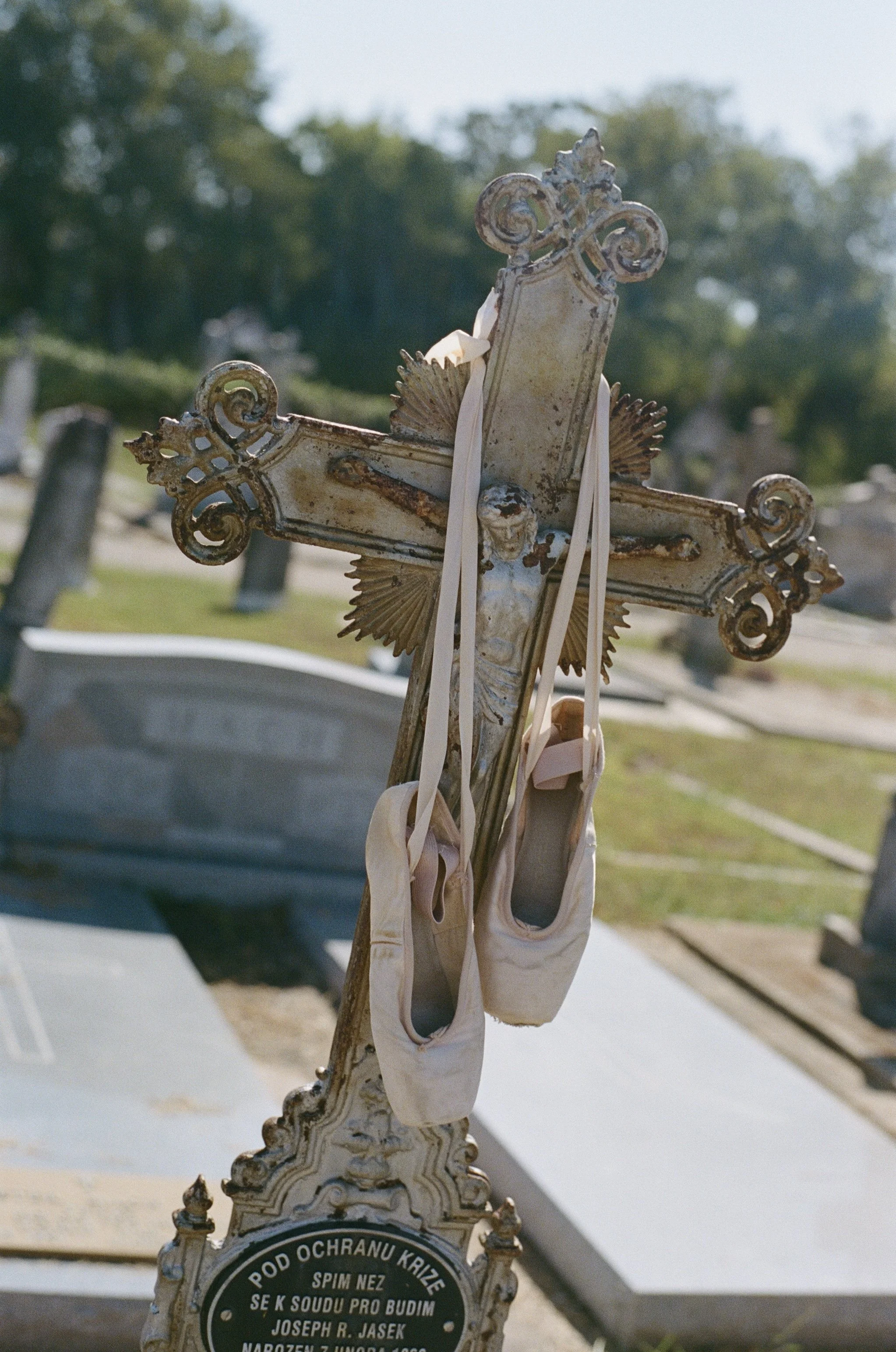 A weathered, ornate metal cross with ballet slippers hanging from it, set in a cemetery with gravestones and trees in the background.