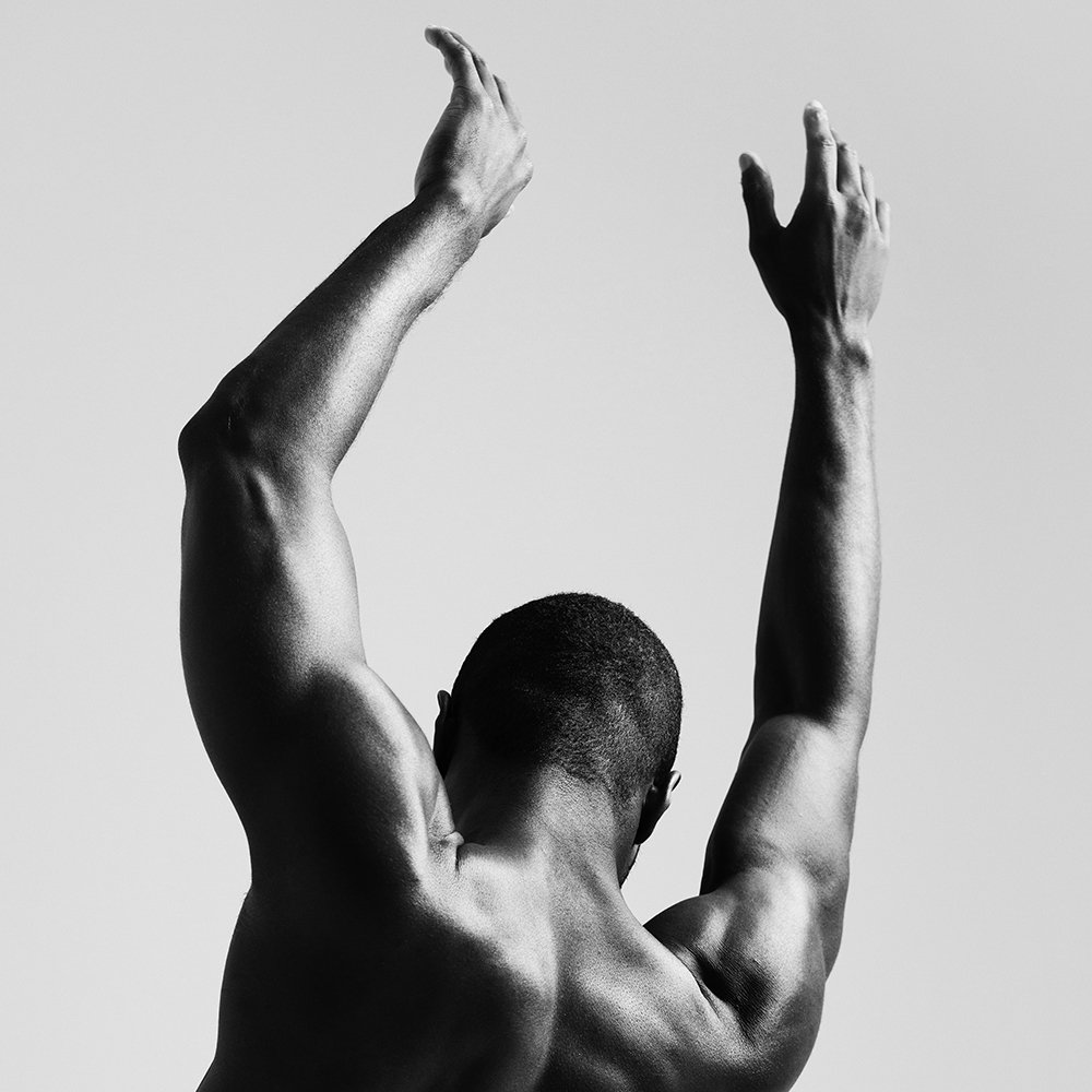 Black and white photo of a person with short hair raising their arms above their head, viewed from the back, showcasing muscular arms and shoulders.