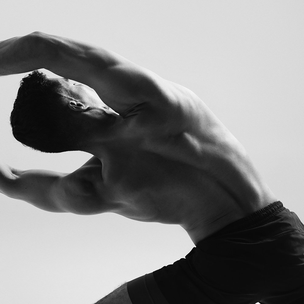 Black and white photo of a shirtless man stretching with arms extended overhead, highlighting muscular back.