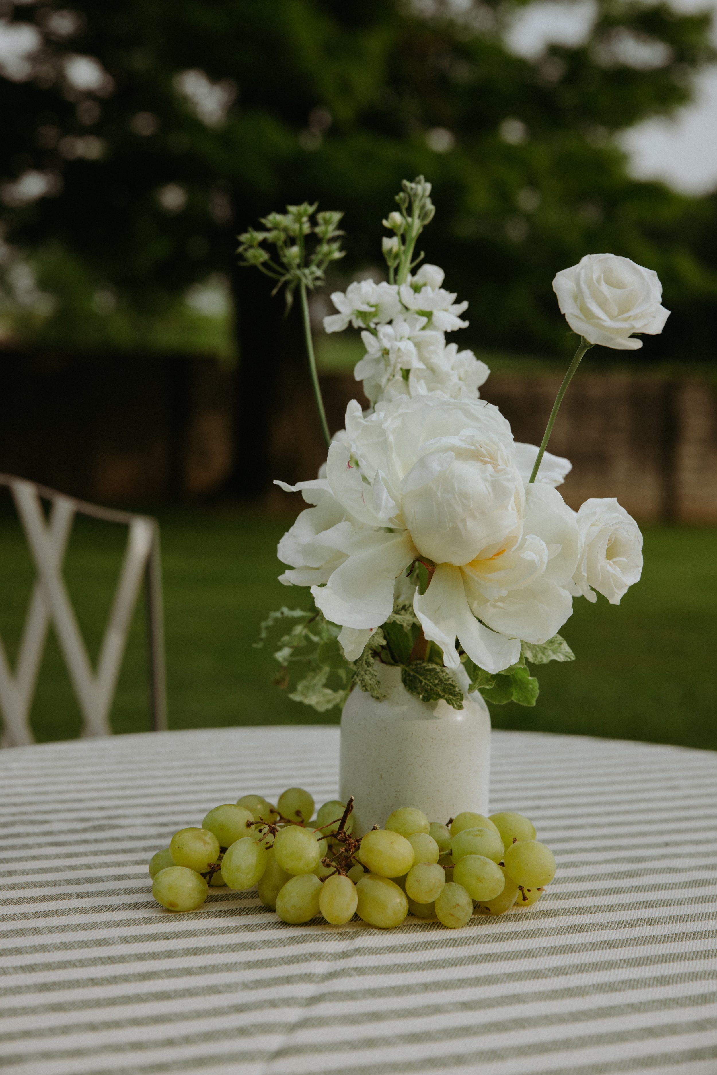 Bud vase arrangement with ivory flowers and grapes on a striped linen tablecloth