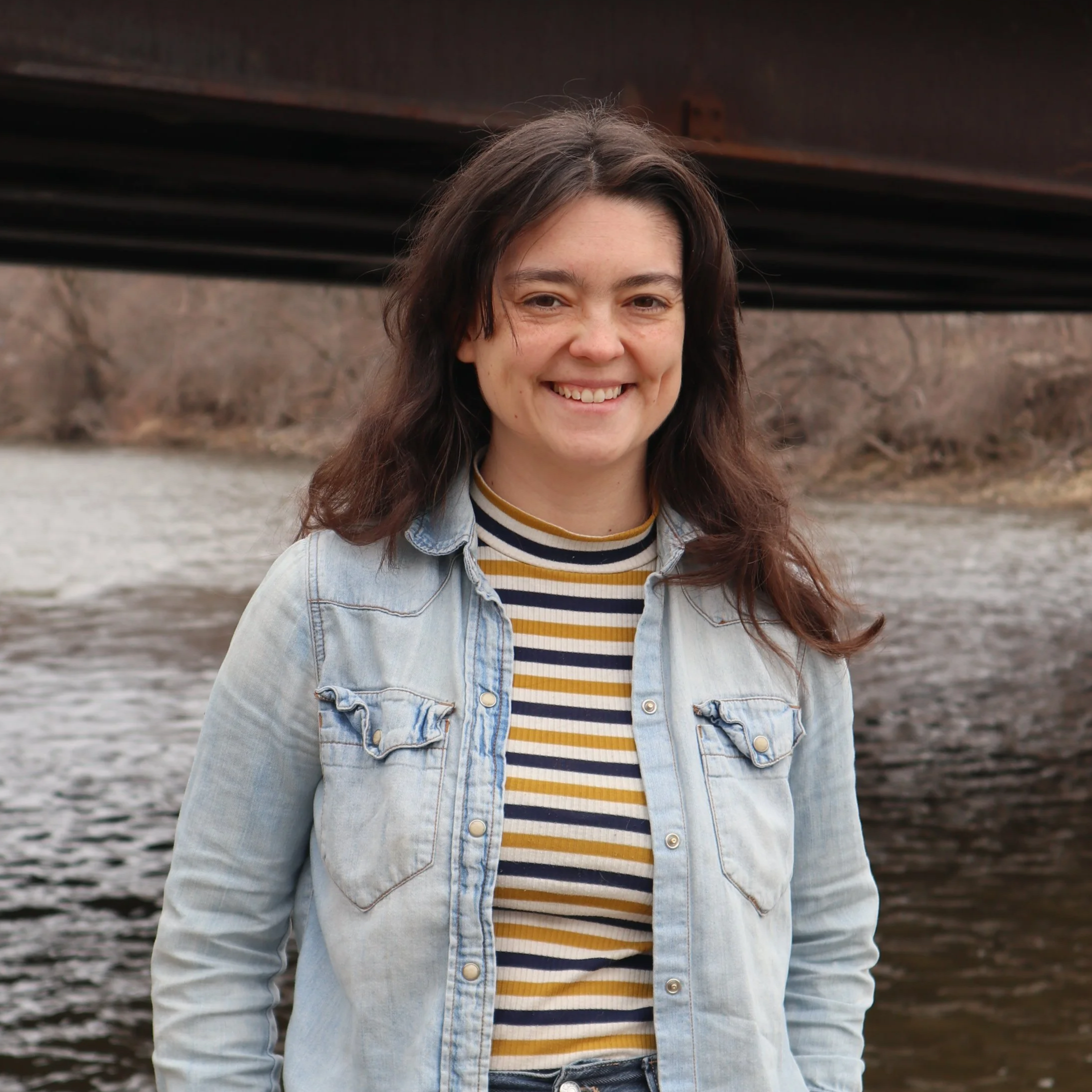 A woman is smiling and standing near a river under a bridge. She wears a denim shirt over a striped top and jeans. The scene is calm and casual.