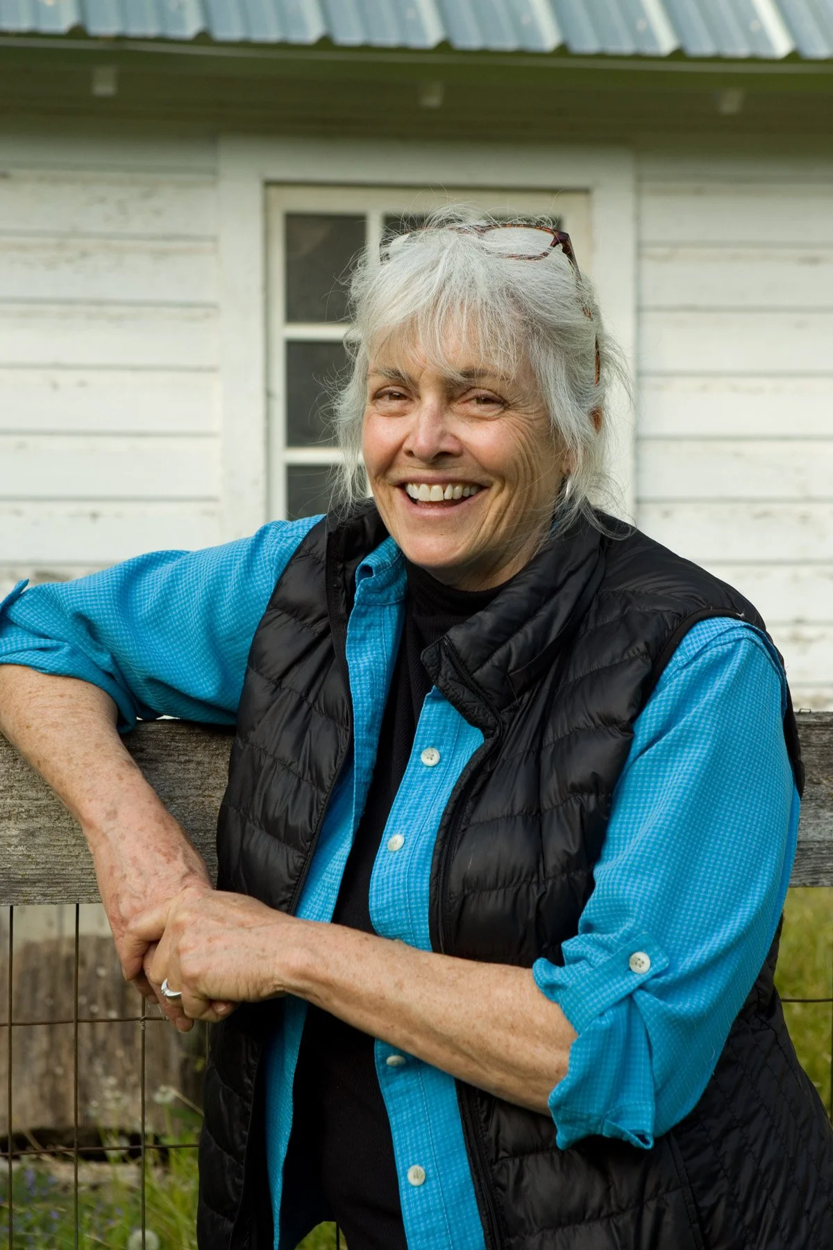 Carrie Jennings, a woman with gray hair and glasses atop her head smiling warmly. She wears a black vest over a blue shirt, leaning on a rustic fence. Background: white wooden house.