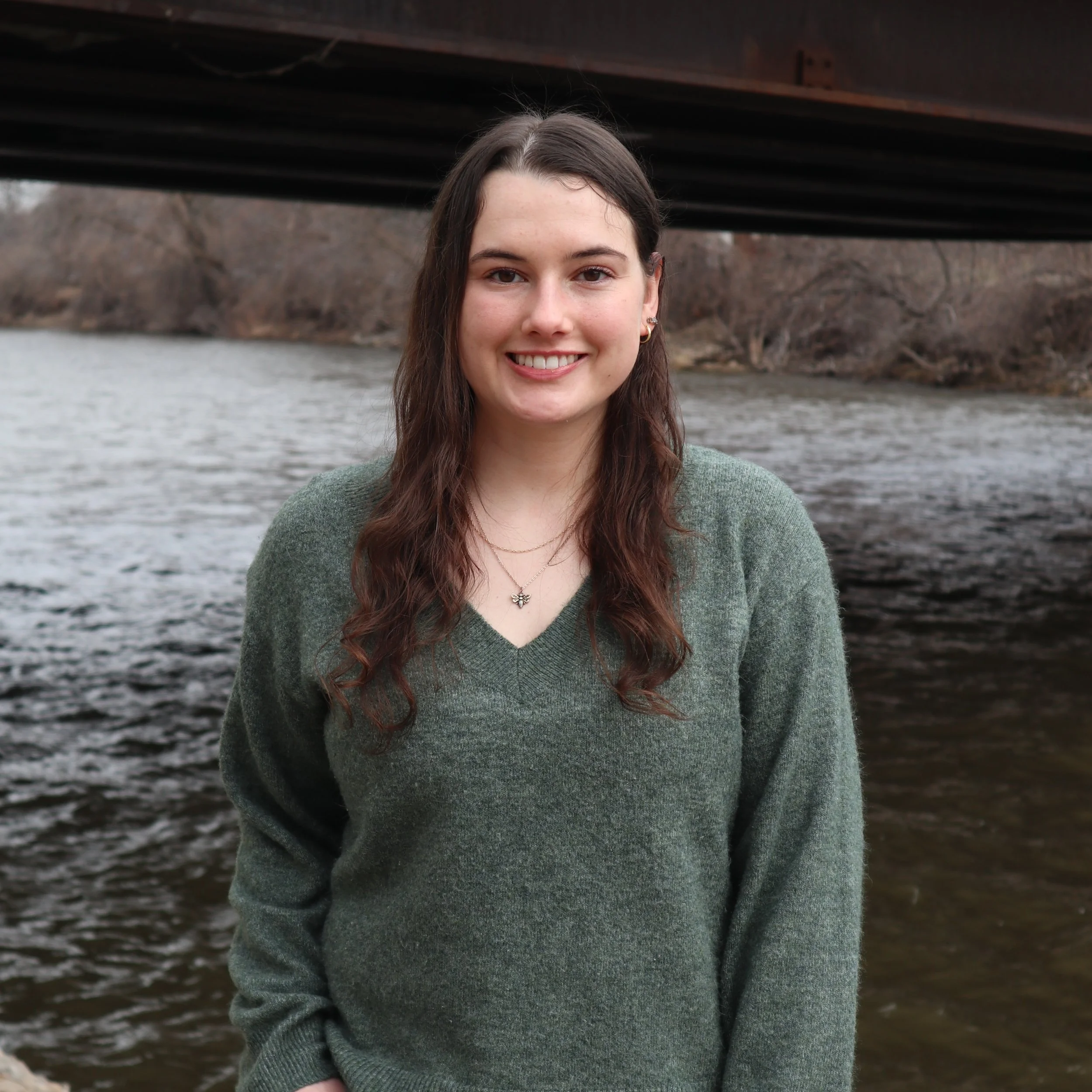 A young woman with long brown hair smiles warmly, standing near a river under a bridge. She wears a green sweater, with a backdrop of bare trees.