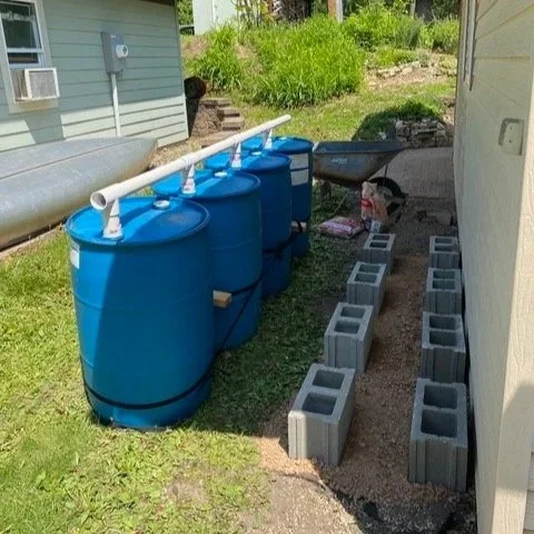  Four blue rain barrels connected by white PVC pipes stand beside a house. Nearby, concrete blocks are neatly arranged on gravel. The setting is a sunny backyard. 