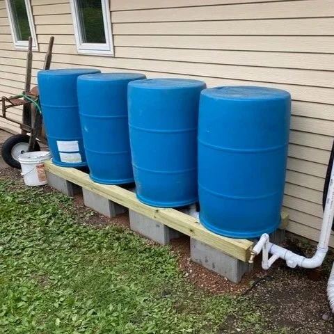  Four blue rain barrels are lined up against a beige house on a wooden platform. Pipes are connected, suggesting a rainwater collection system. 