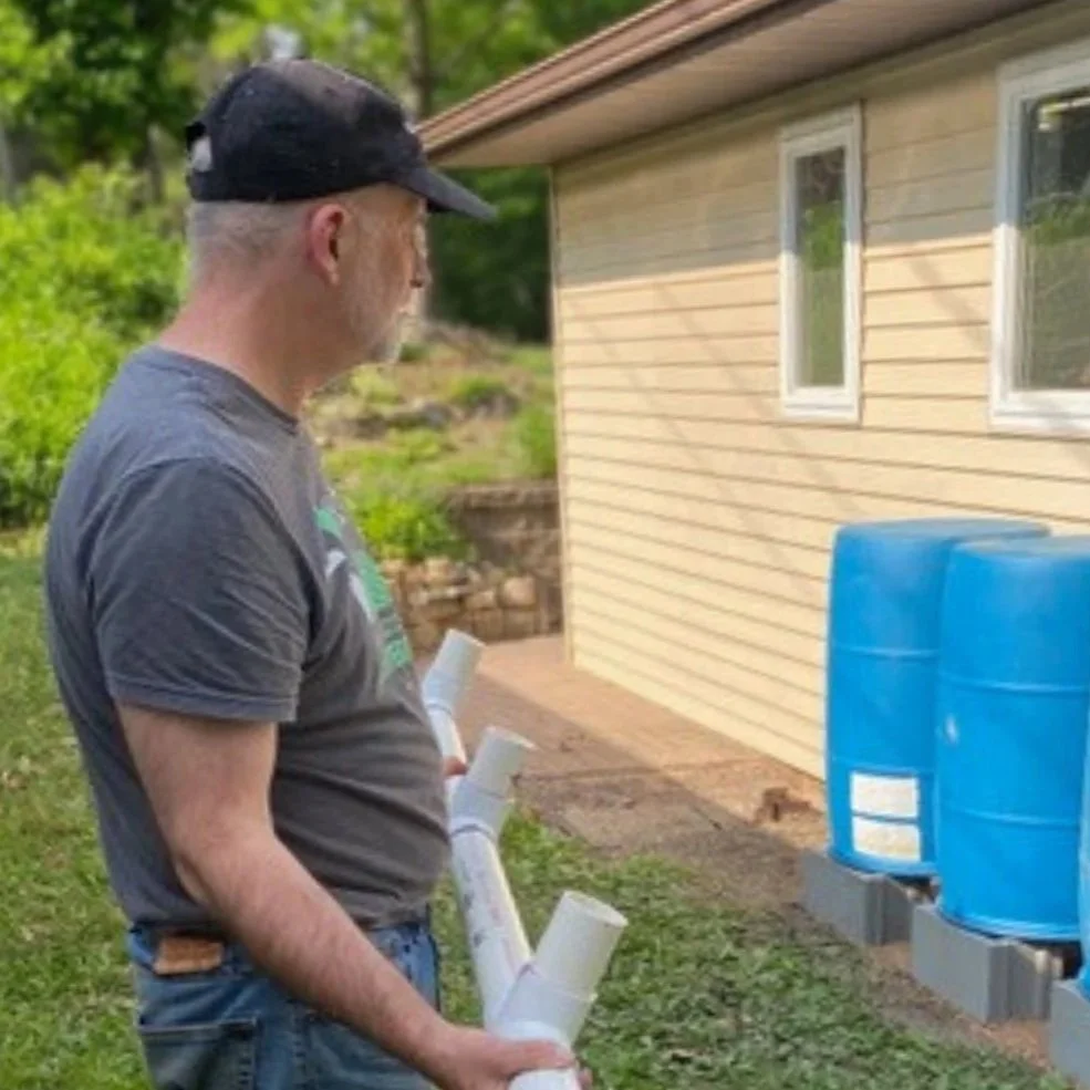  A man in a cap observes blue rain barrels beside a beige house. He's holding PVC pipes, indicating a rainwater harvesting setup. Greenery surrounds the scene. 