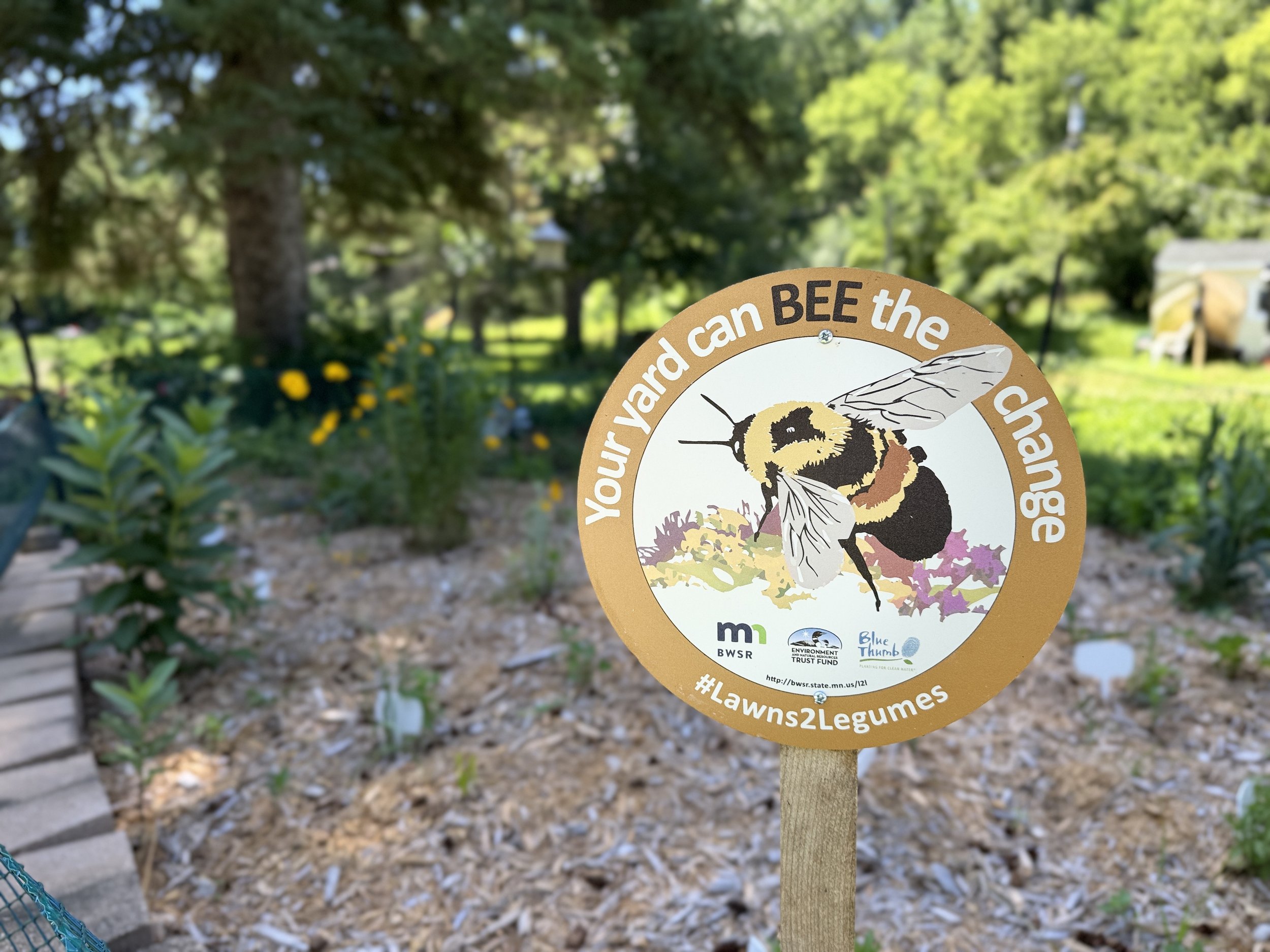 Sign in a garden reads, "Your yard can BEE the change," featuring a bee illustration. Background shows green foliage, creating a serene nature scene.