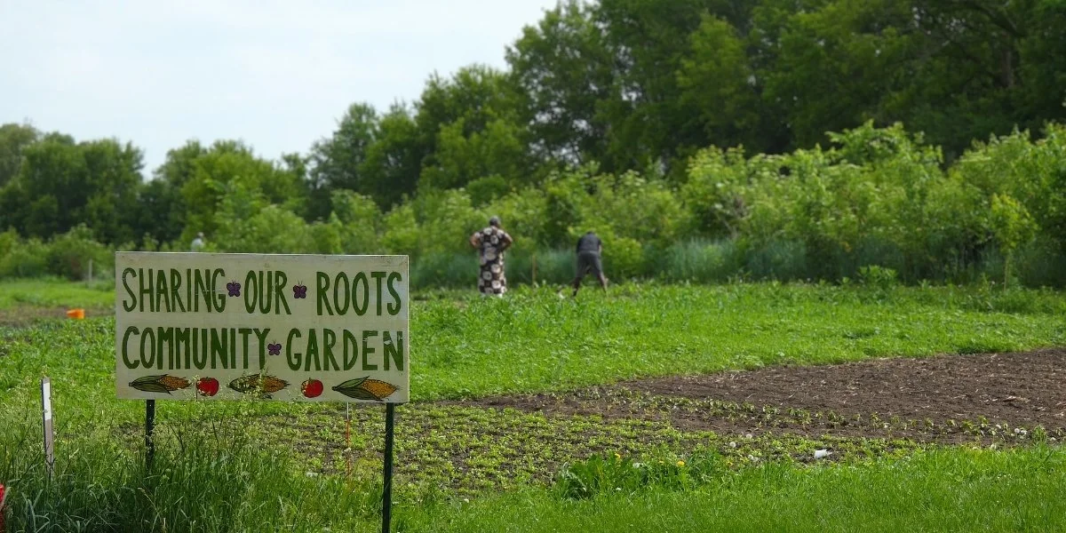 A sign reading "Sharing Our Roots Community Garden" stands in a green field.