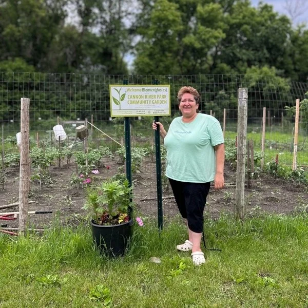 A woman is standing next to a garden sign, with blooming flowers and greenery in the background.