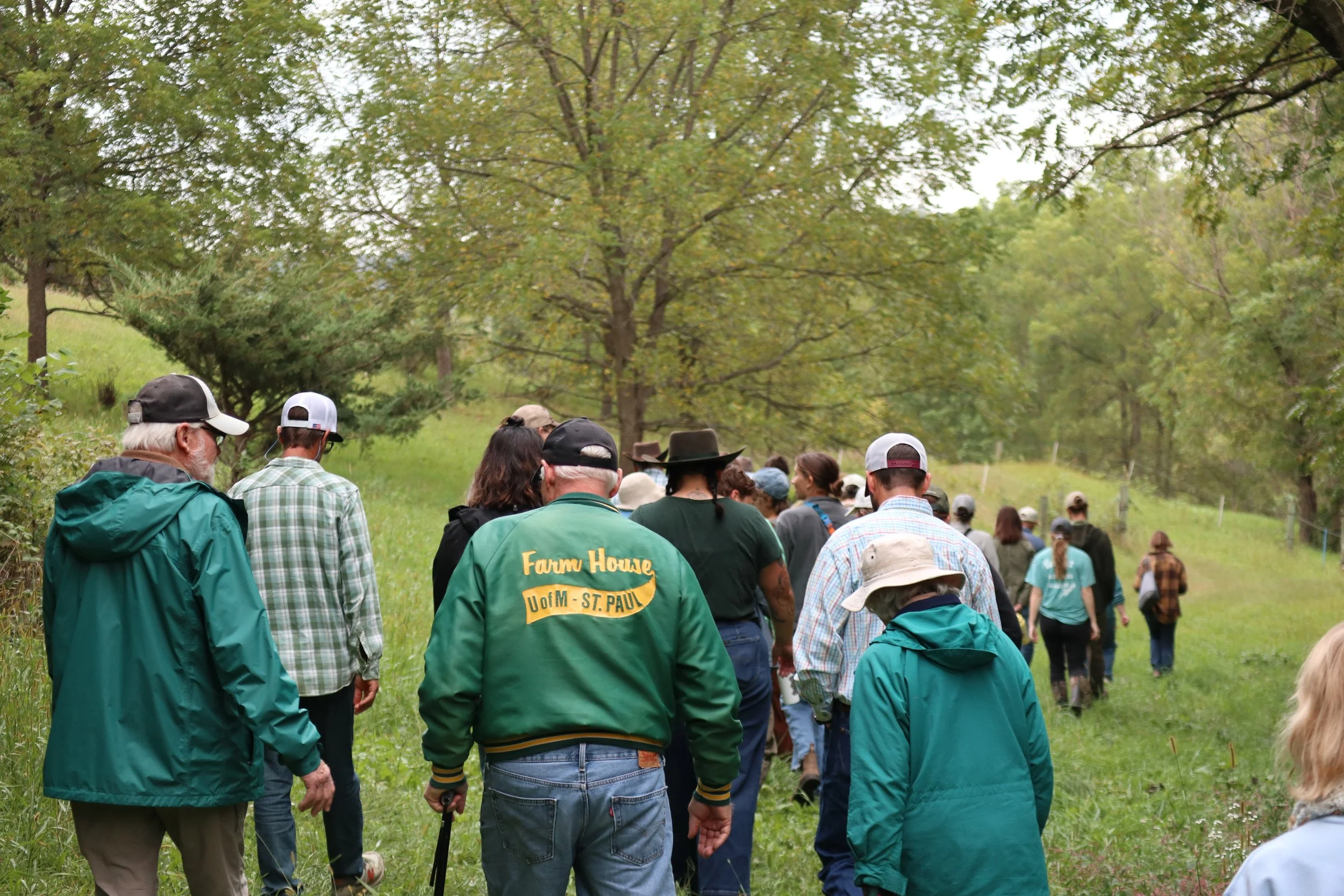A group of people walks through a forested trail, wearing casual outdoor clothing. The scene is lively, surrounded by green trees and lush grass.