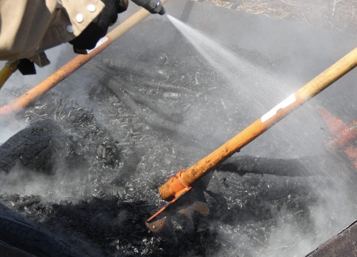 A person sprays water on smoldering charred debris (biochar), while orange-handled tools lie across the blacked surface, emitting wisps of smoke.