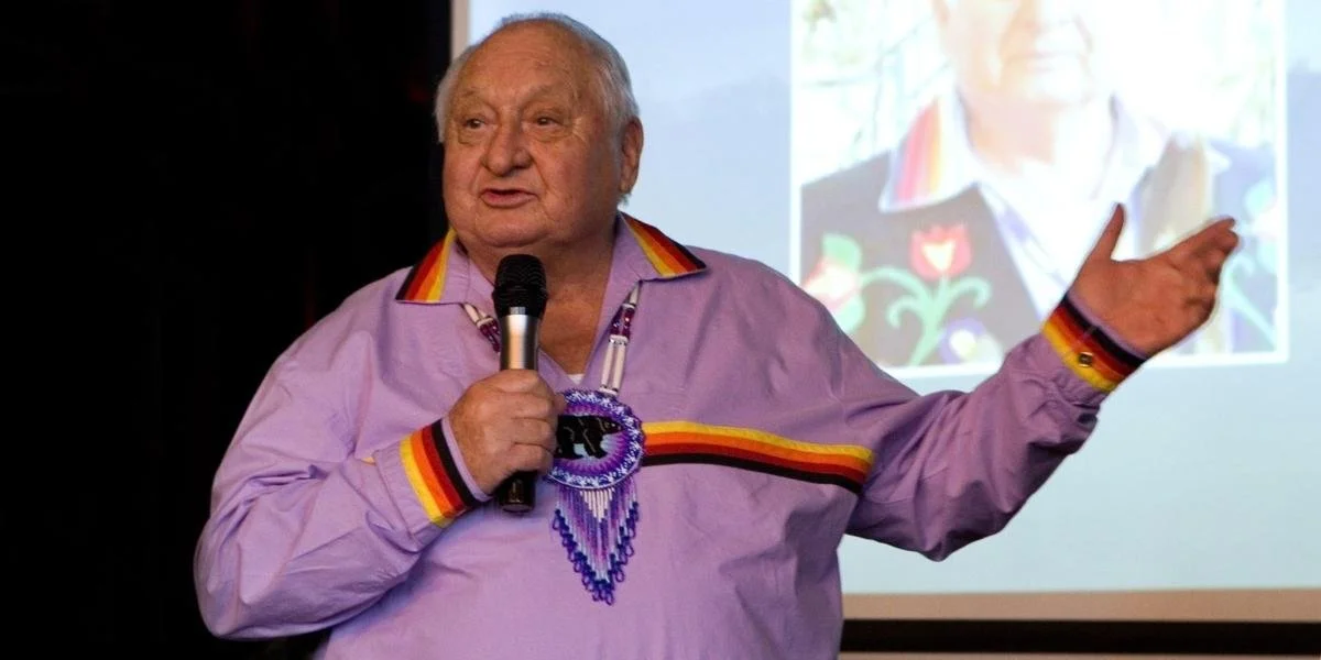 Elderly man in a lavender shirt with colorful trim speaks into a microphone on stage, gesturing warmly. A serene, engaged expression on his face.