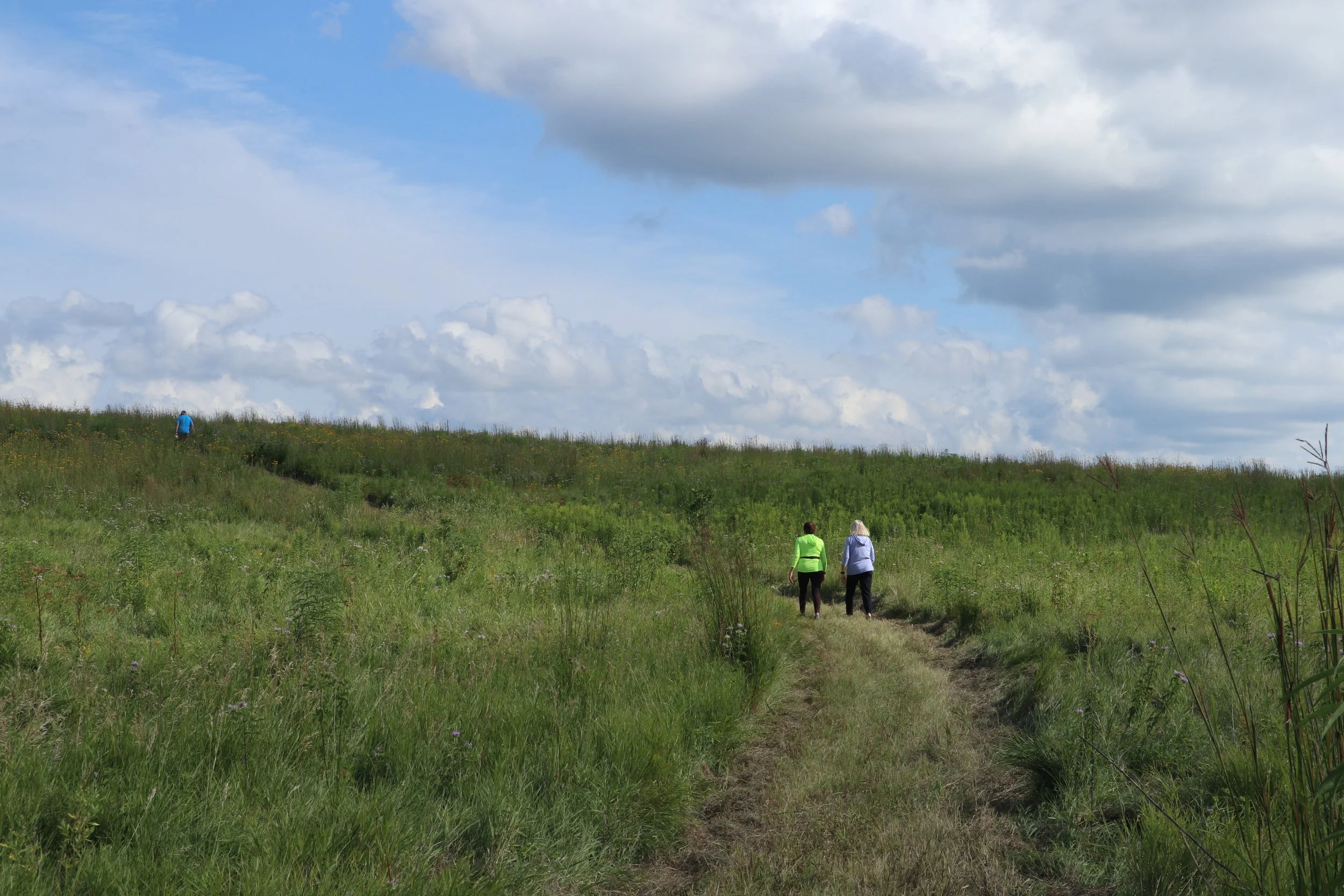 Two people in bright jackets walk along a grassy path under a partly cloudy sky. The open field and distant horizon create a calm, expansive feel.