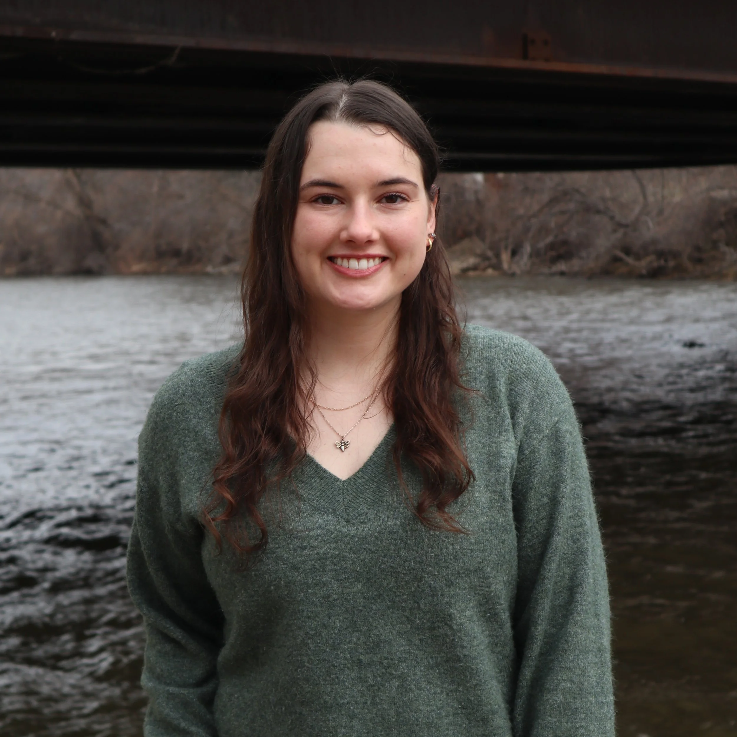 A young woman with long brown hair smiles warmly, standing near a river under a bridge. She wears a green sweater, with a backdrop of bare trees.