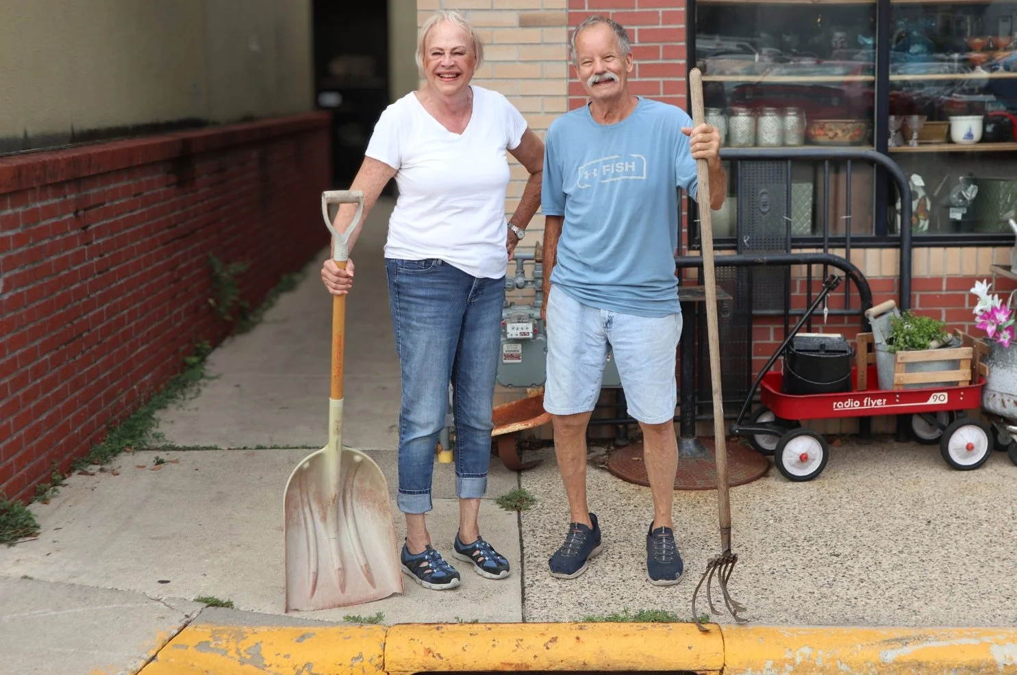 A man and a woman holding tools in front of a local business and storm drain.