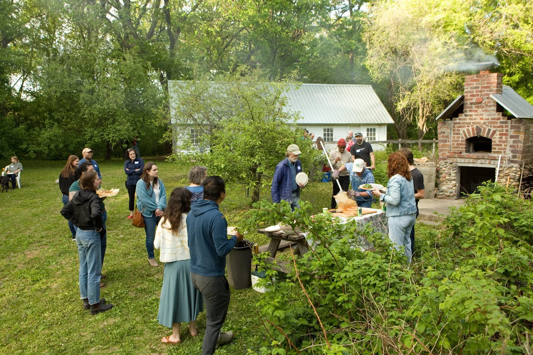 A group of people standing around in a backyard setting, on a farm, eating pizza from a wood fired oven.