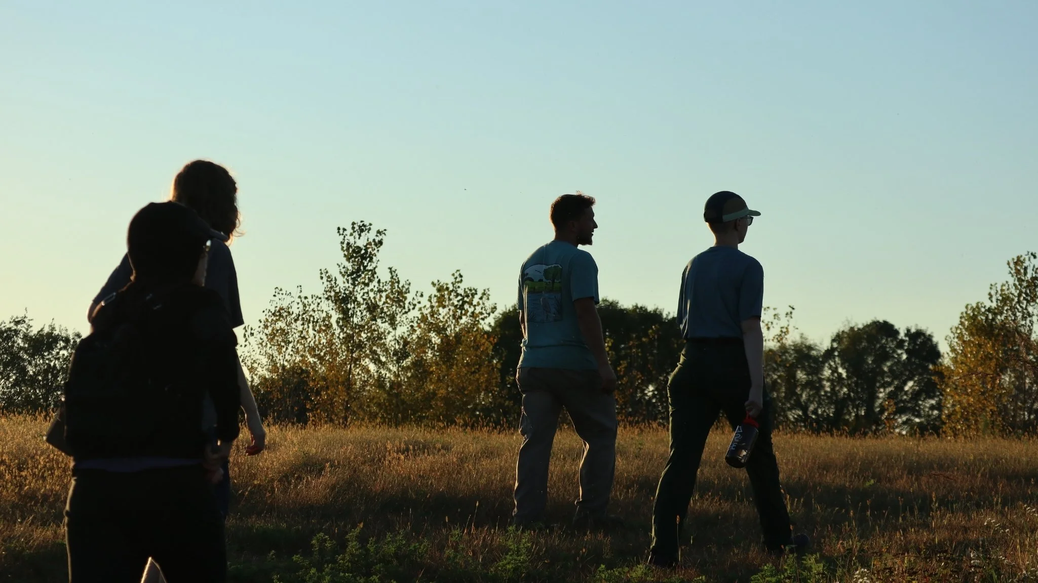 A group of people walking up a grassy hill shadowed by the sunset with blue skies and greenery.