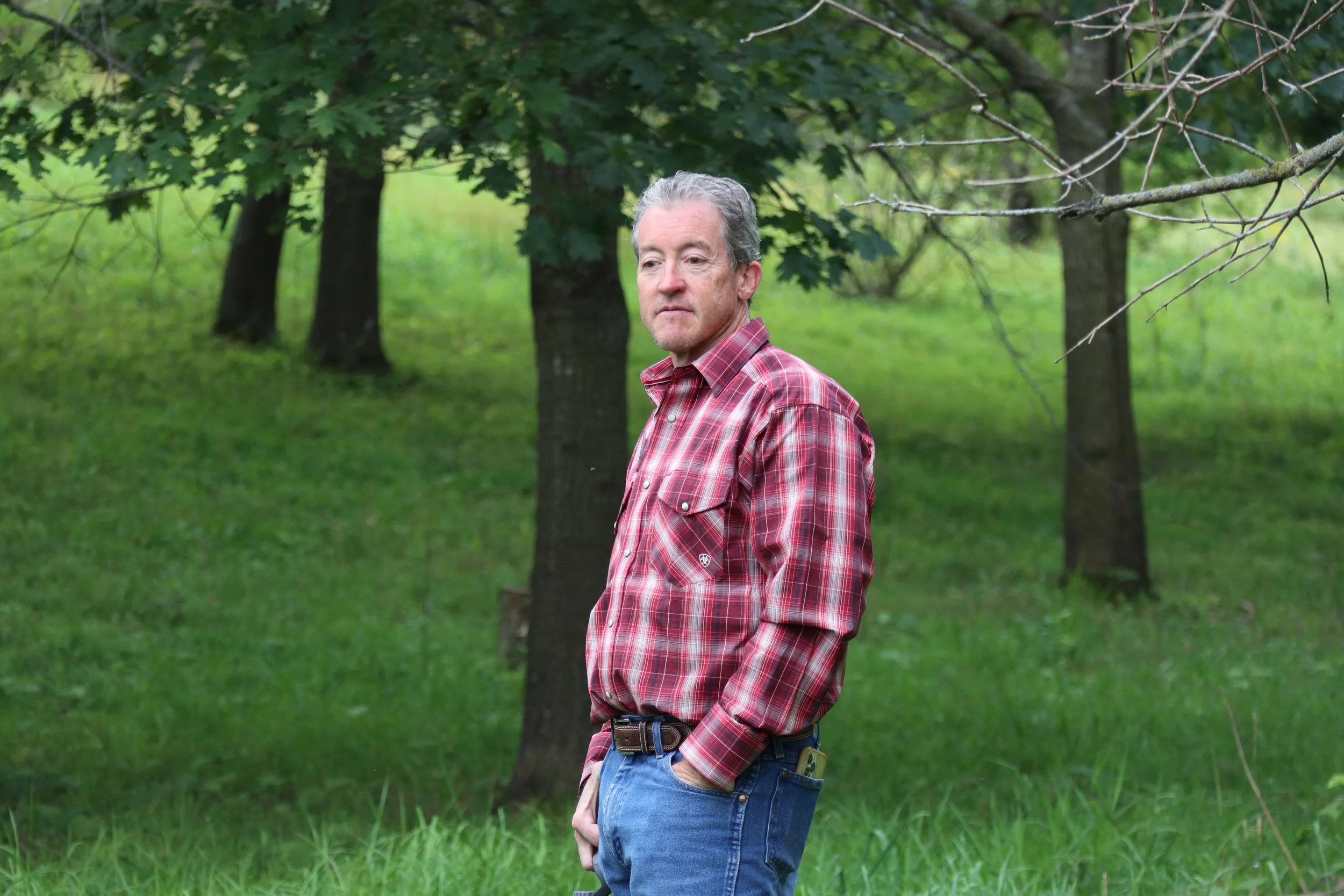 Todd Churchill, a man in a red plaid shirt stands thoughtfully in a lush, green park. Trees surround him, creating a peaceful and contemplative atmosphere.