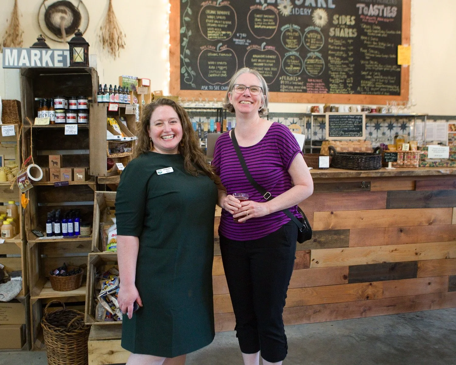 Two women smiling in a cozy market. The woman on the left wears a green dress, and the one on the right wears a purple striped top. Wooden shelves with products and a large menu chalkboard are in the background. Warm, inviting atmosphere.