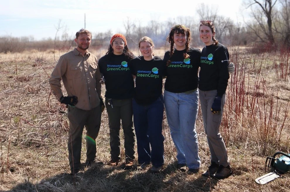 A group of five people in fieldwork attire stand closely together, smiling. They wear "Minnesota GreenCorps" shirts, set against a background of dry grass and leafless trees, suggesting early spring. The mood is cheerful and collaborative.