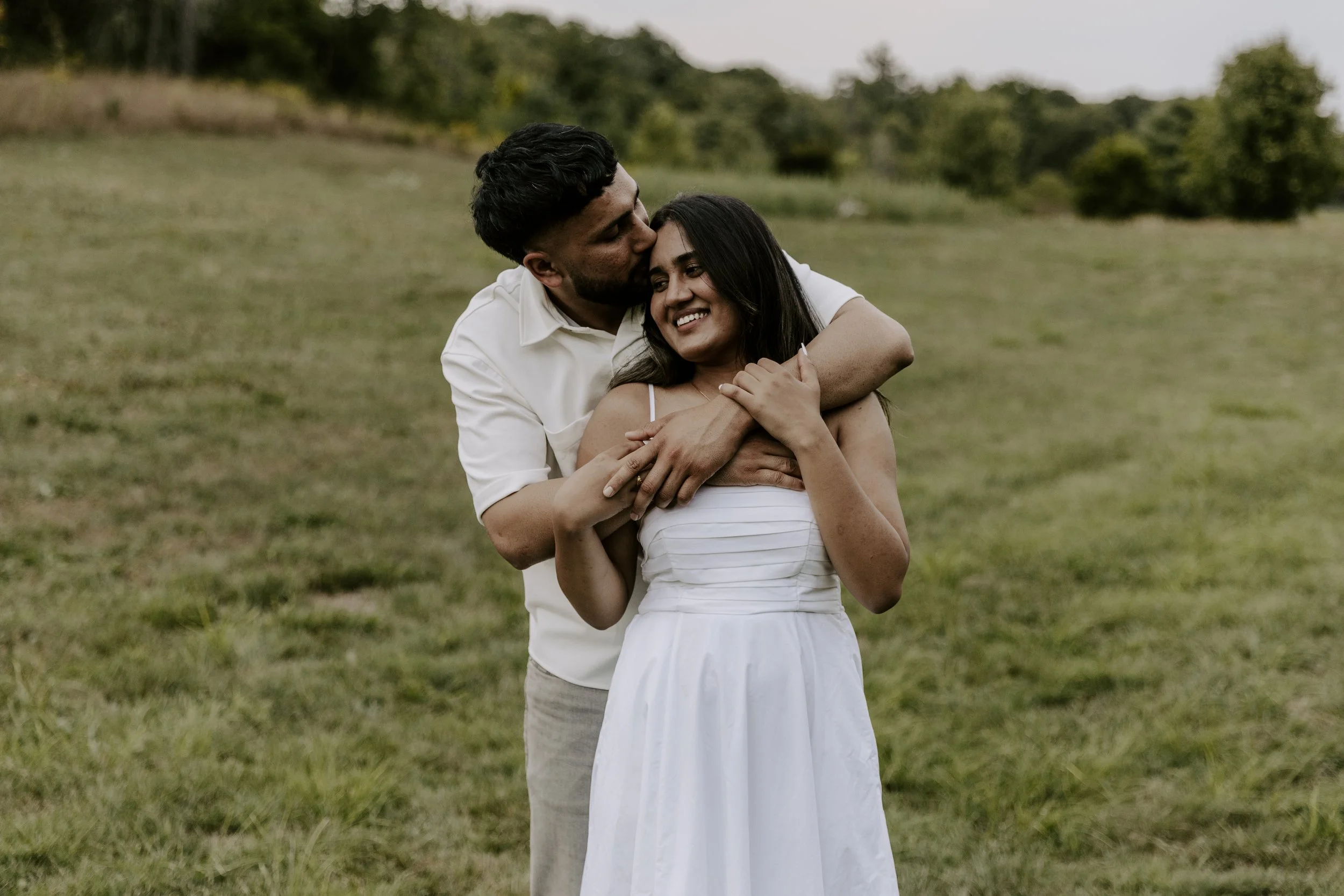 A man and woman embrace outdoors in a grassy field; the man kisses the woman's forehead as she smiles happily with her eyes closed during their engagement photography package in Ontario.