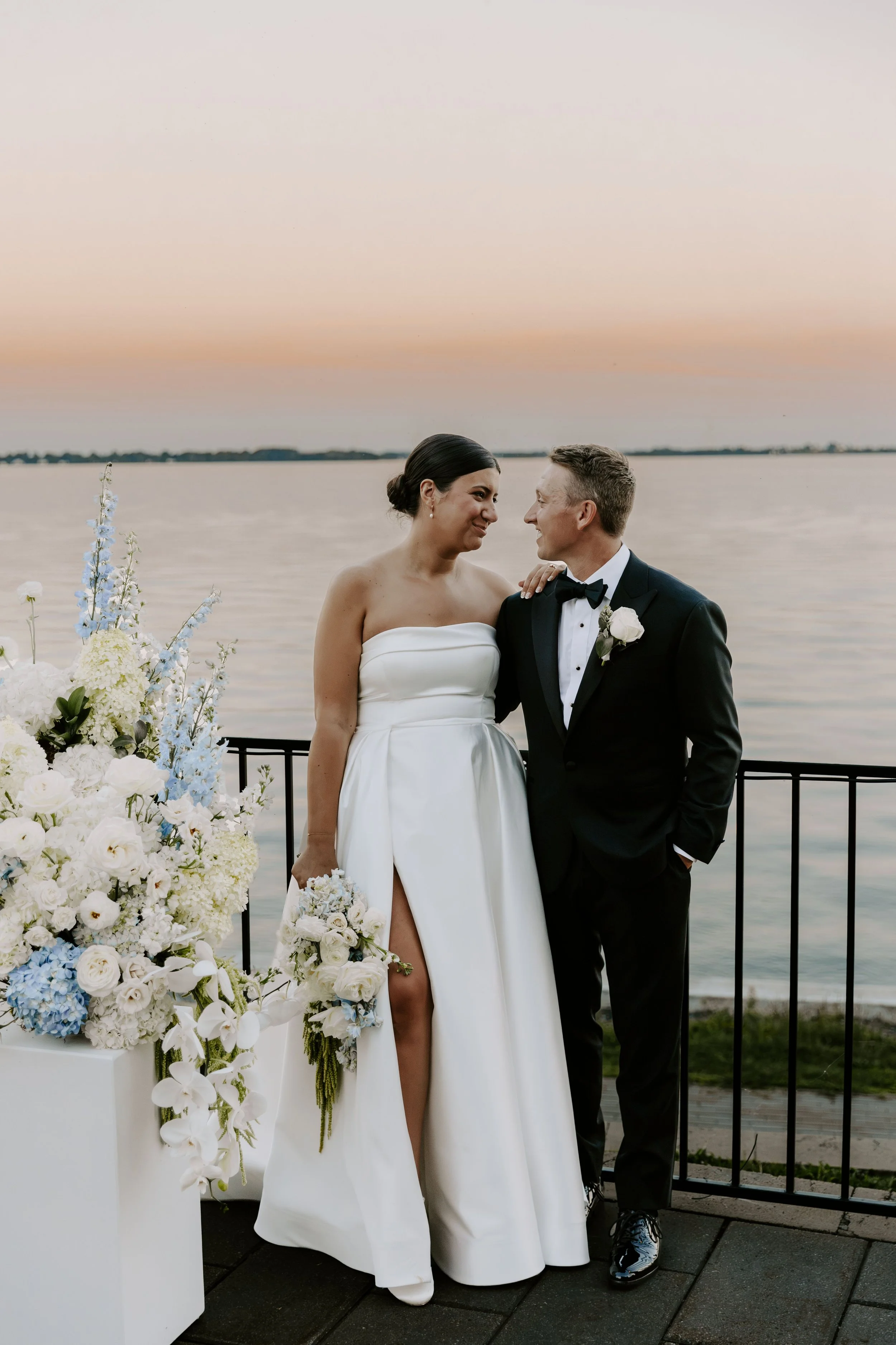 A bride and groom standing close to each other near a body of water at sunset, smiling and looking into each other's eyes. The bride is wearing a strapless white wedding dress with a high slit, holding a bouquet of white and blue flowers. The groom is in a black tuxedo with a bow tie and boutonniere. There are white flower arrangements in front of them.