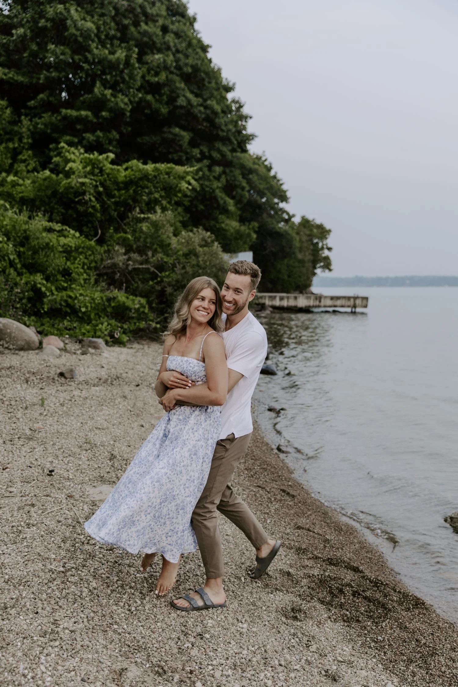 A happy couple hugging and smiling on a rocky beach near a body of water, with a wooded area behind them and a pier extending into the water during their couples photography package in Ottawa Ontario.