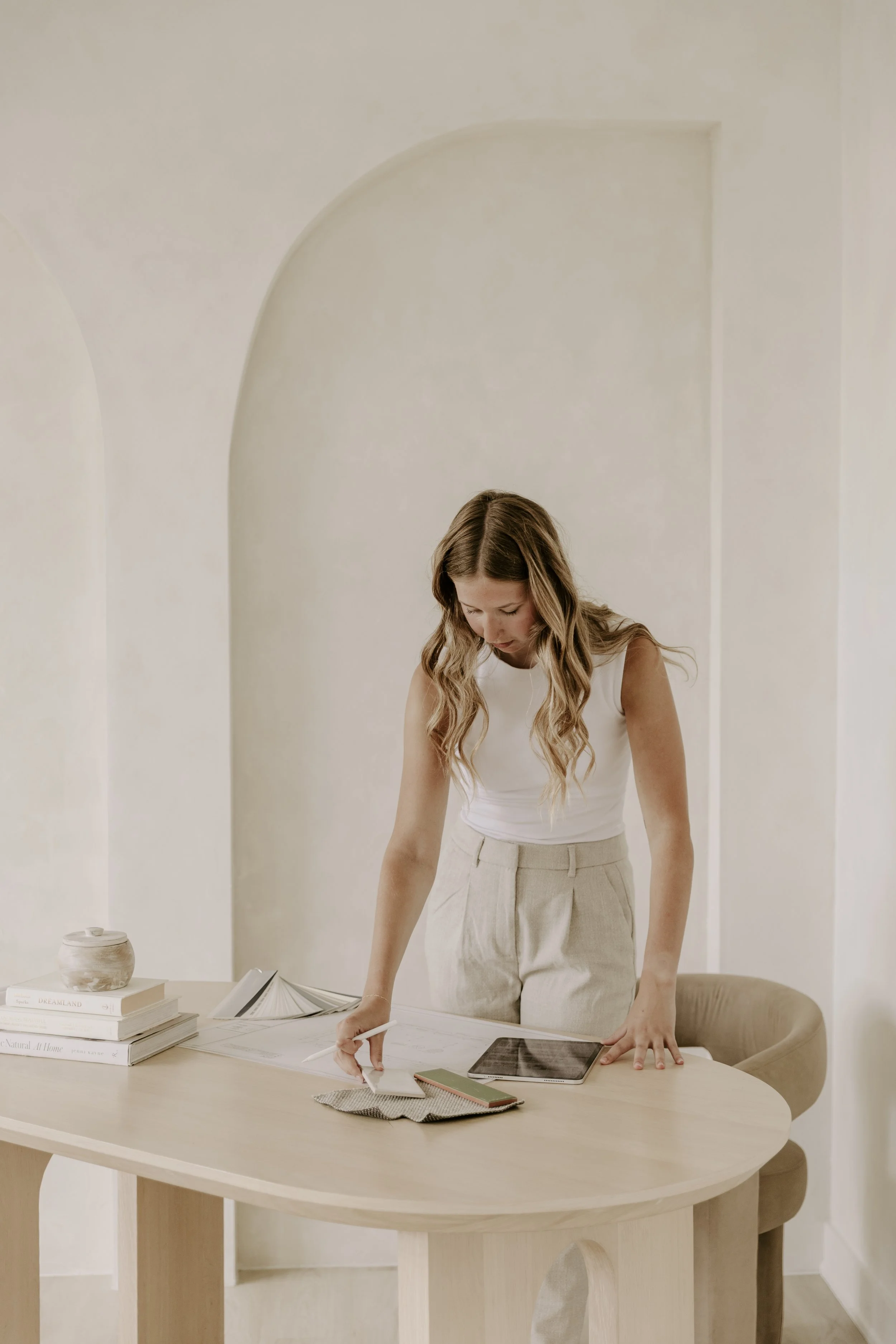 Woman stands at a table, looking down at documents, with a tablet, notebook, and books on the table. The room has white walls and a minimal decor for branding photography package in Ontario.