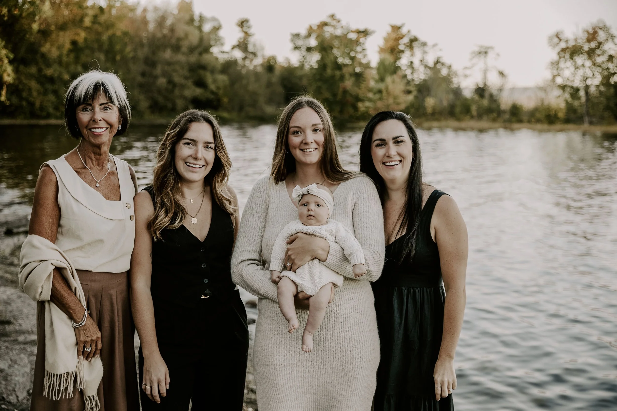 Group of five women and a baby standing by a body of water with trees in the background, during the evening during extended family photography package in Eastern Ontario.