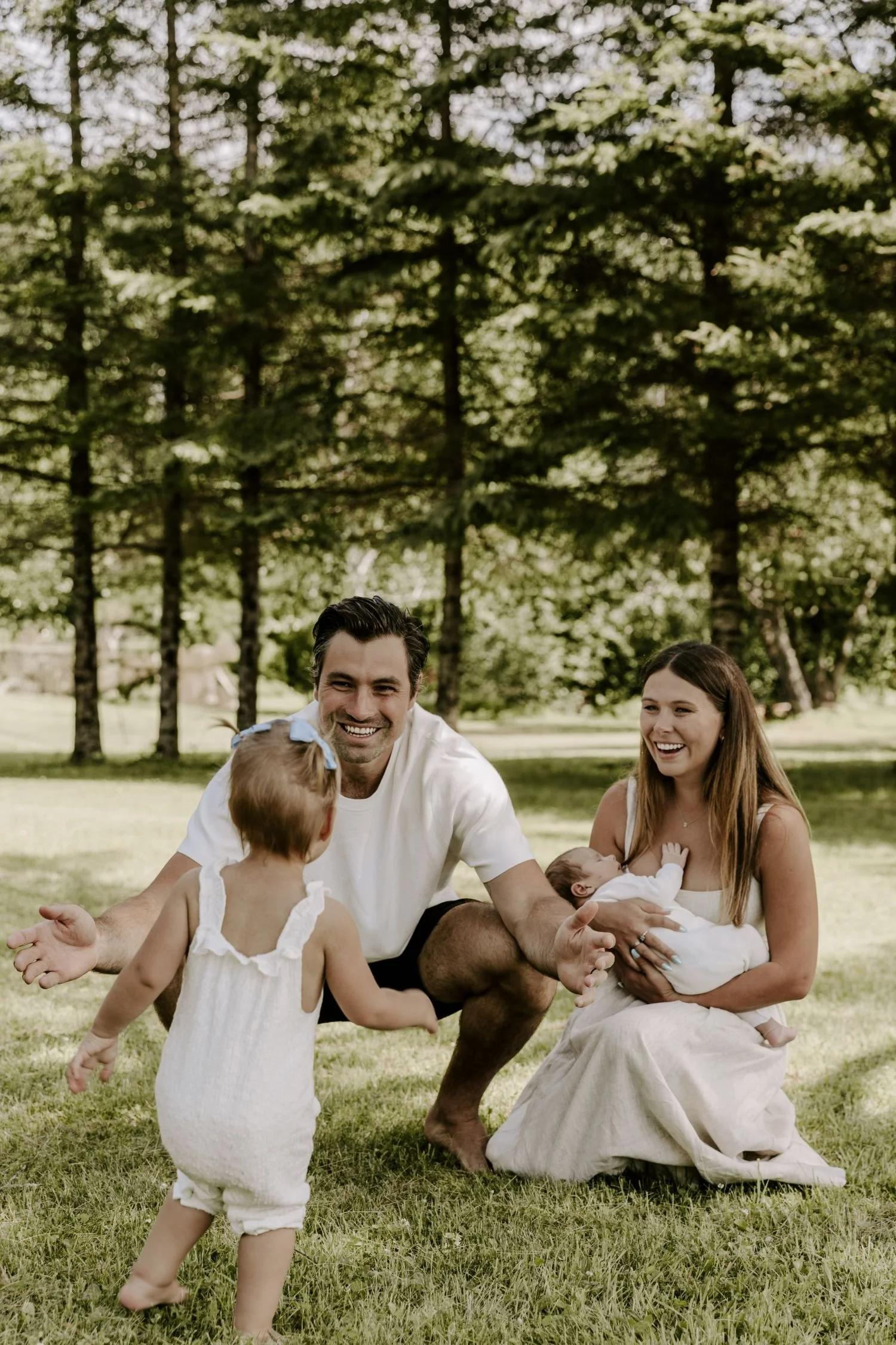 A family of five, including a man, woman, two young children, and a baby, sitting and playing on a grassy field with trees in the background, enjoying an outdoor moment for family photography package in Eastern Ontario.