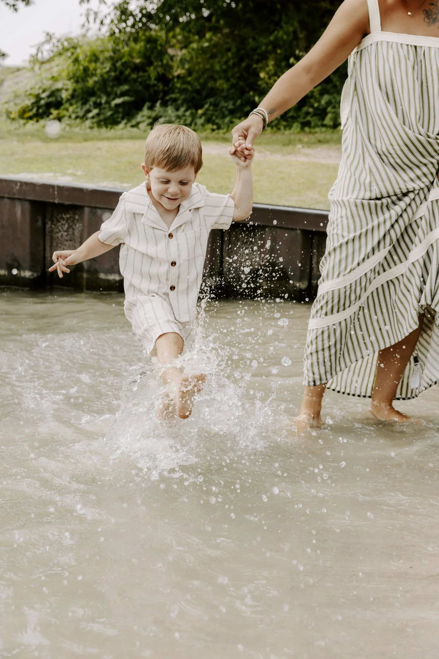 A young boy in striped pajamas playing and walking in shallow water while holding hands with an adult woman, possibly his mother, in a striped dress, outdoors near a wooden structure and greenery for family photography package in Ontario.