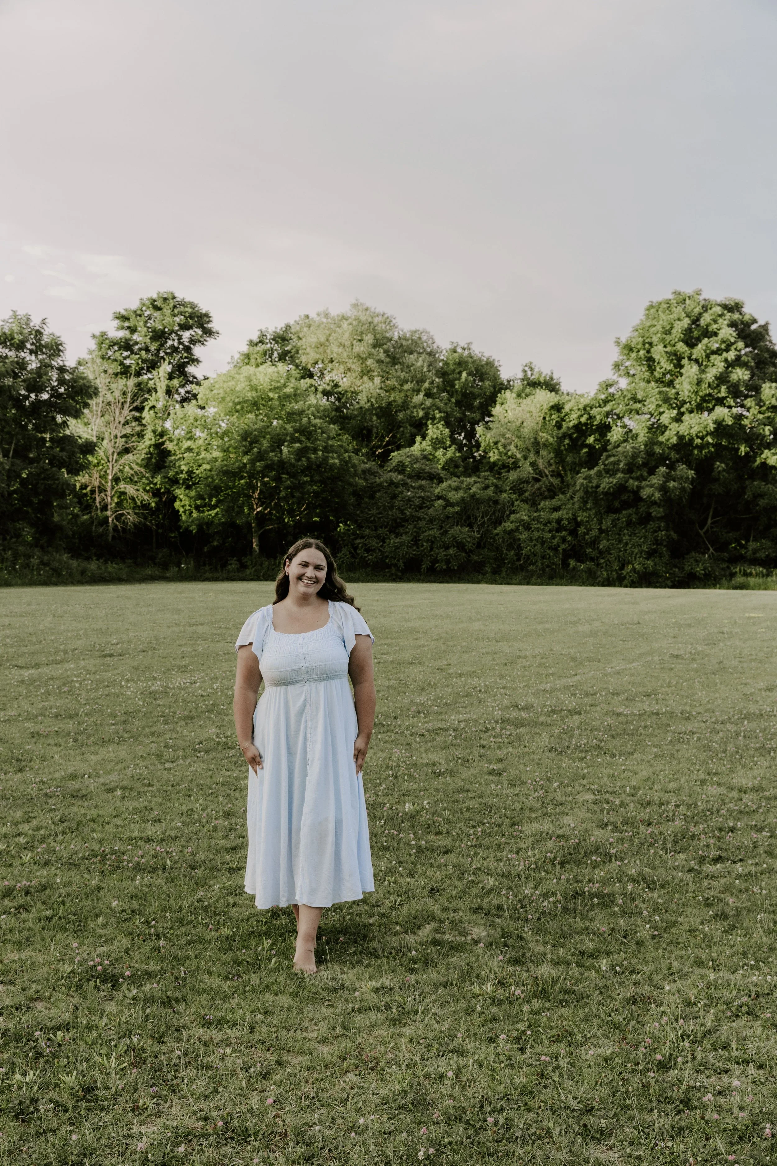 A woman in a white dress standing barefoot on a grassy field with trees in the background for wedding, elopement, couples and family photographer in Eastern Ontario.