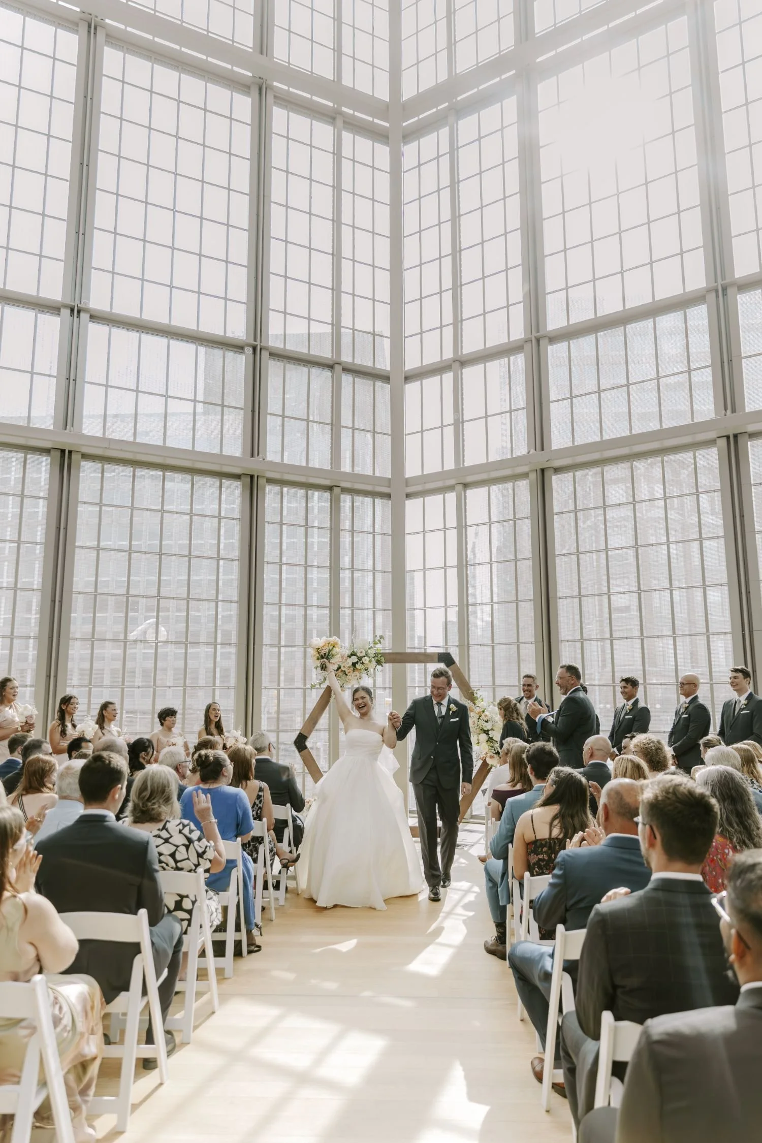Indoor wedding ceremony with a bride and groom walking down the aisle, surrounded by guests seated on both sides, large glass windows in the background, and a floral arrangement visible for wedding photography package in Ottawa area Ontario.