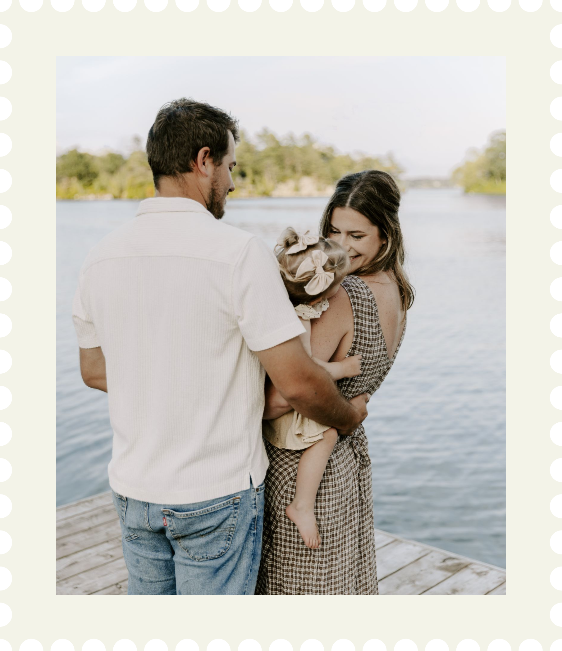 A family of three standing on a dock by a lake, smiling and embracing each other during daytime for family photography package in Eastern Ontario.