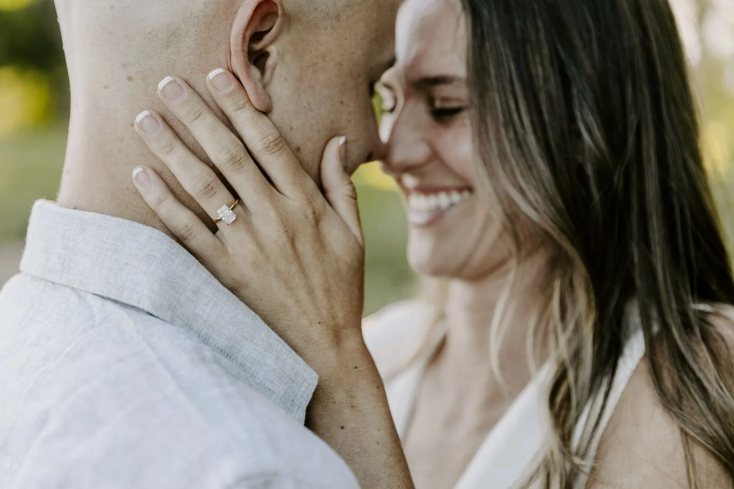 Close-up of a smiling woman with long brown hair touching the face of a man with a bald head, holding her hand near his cheek, showing an engagement ring with a large rectangular diamond for their engagement photography session in Ontario.