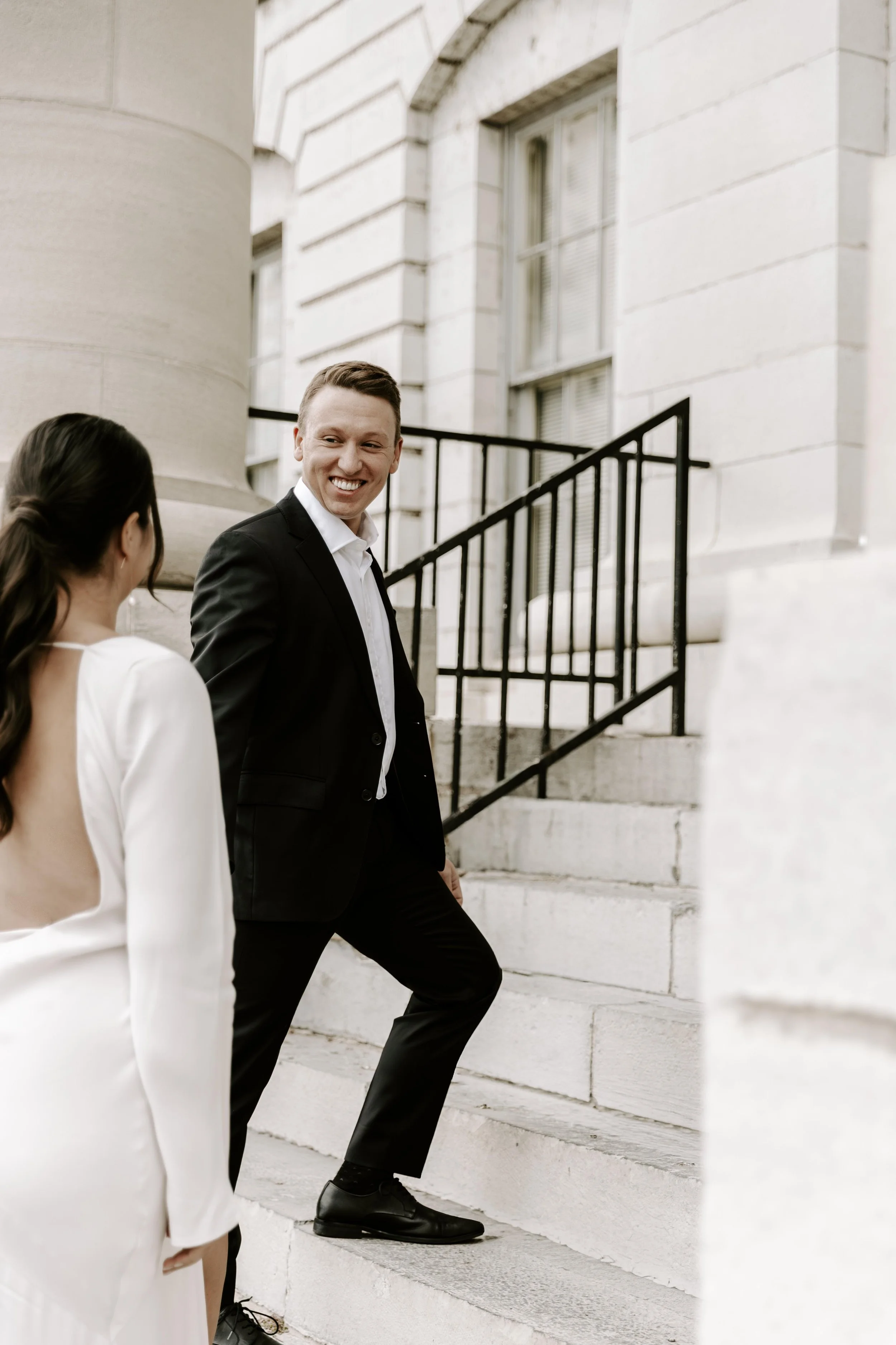 A man in a black suit and woman in a white blazer engaging in conversation outside during their courthouse elopement package in Brockville Ontario.