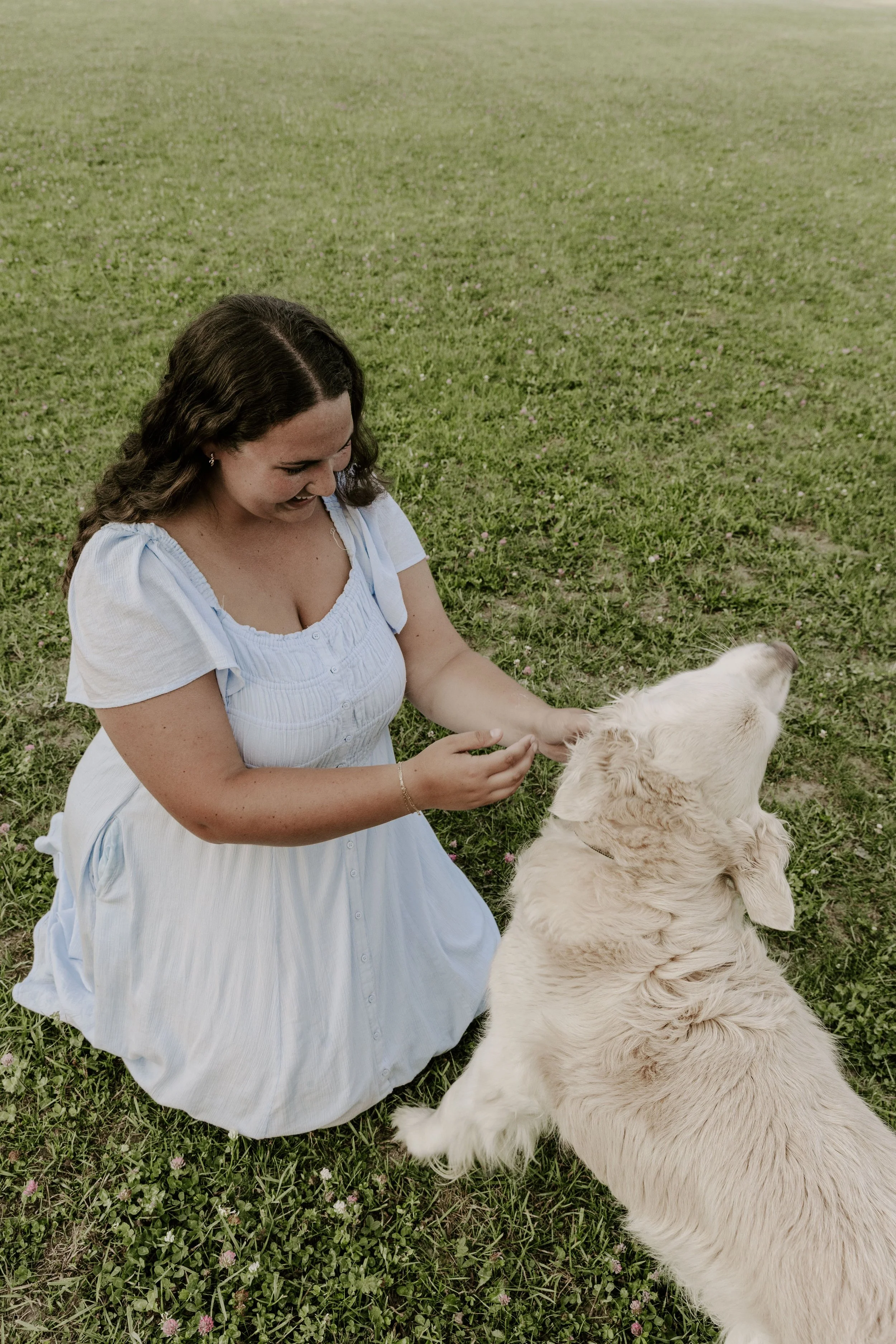 A family, weddings, elopements and couples photographer. She is wearing a blue dress, has dark brown hair and is petting a light coloured golden retreiver.
