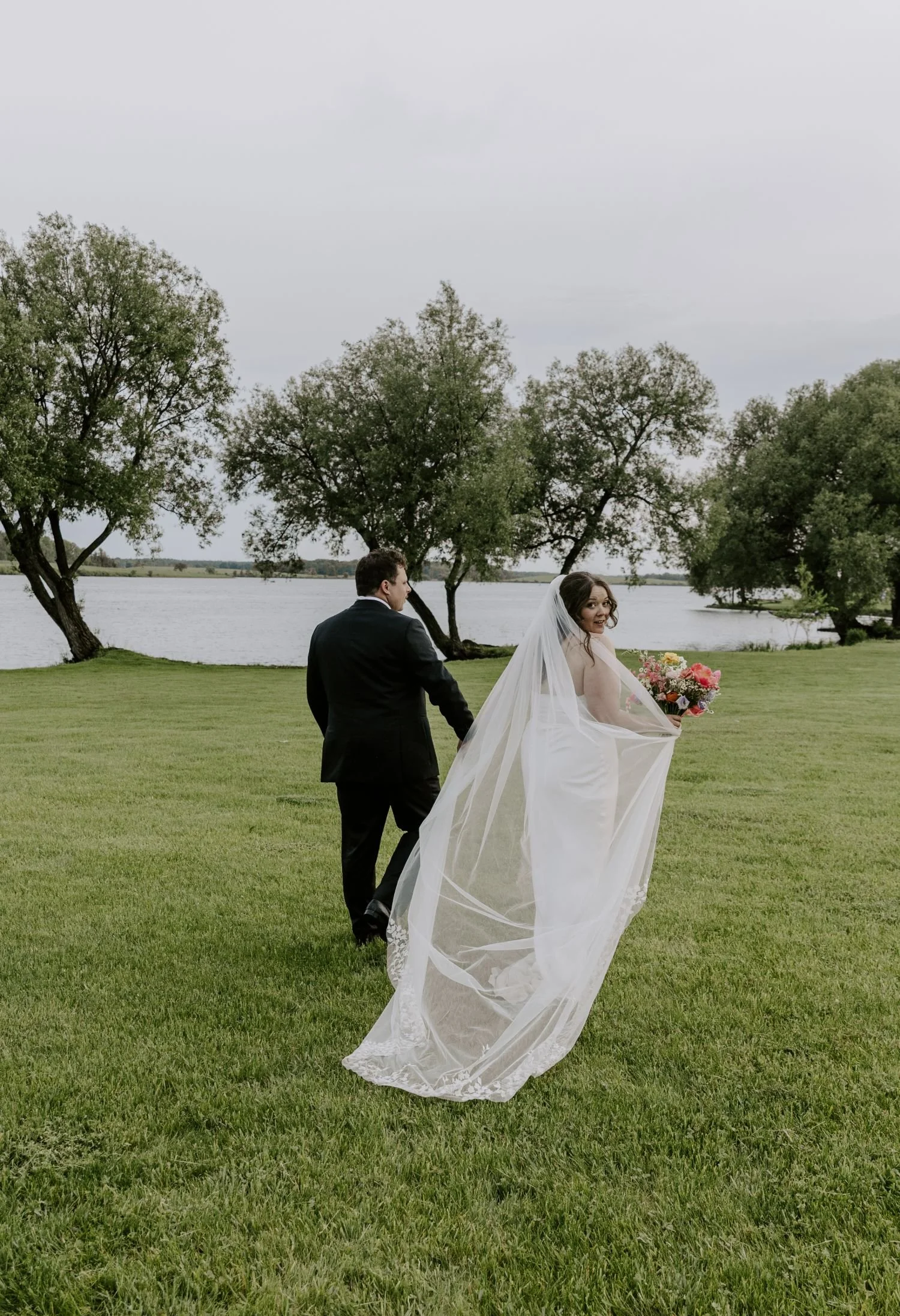 A bride and groom holding hands in an outdoor setting by a lake, with trees in the background, on a cloudy day for their wedding photos in Brockville Ontario.