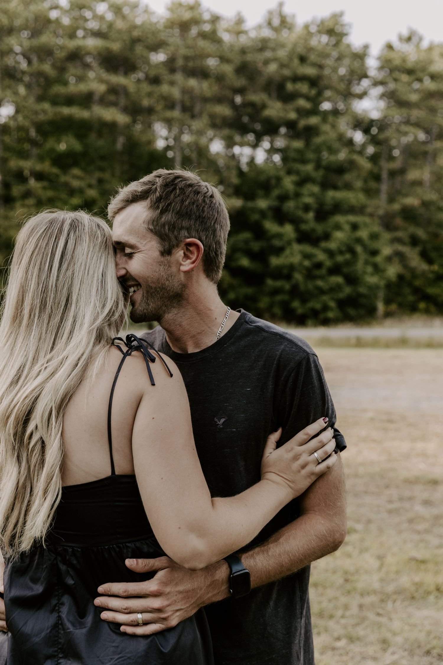 A man and a woman are embracing and smiling outdoors near trees, with a woman having long blonde hair and wearing a black sleeveless top and a man with short brown hair, a beard, and wearing a black t-shirt for couples photography package in Ontario.