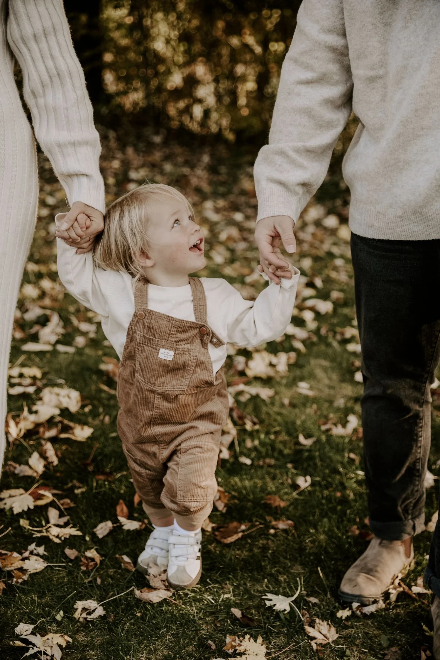 A young child holding hands with two adults outdoors among fallen autumn leaves, with the focus on the child's joyful face for family photography packages in Brockville Ontario