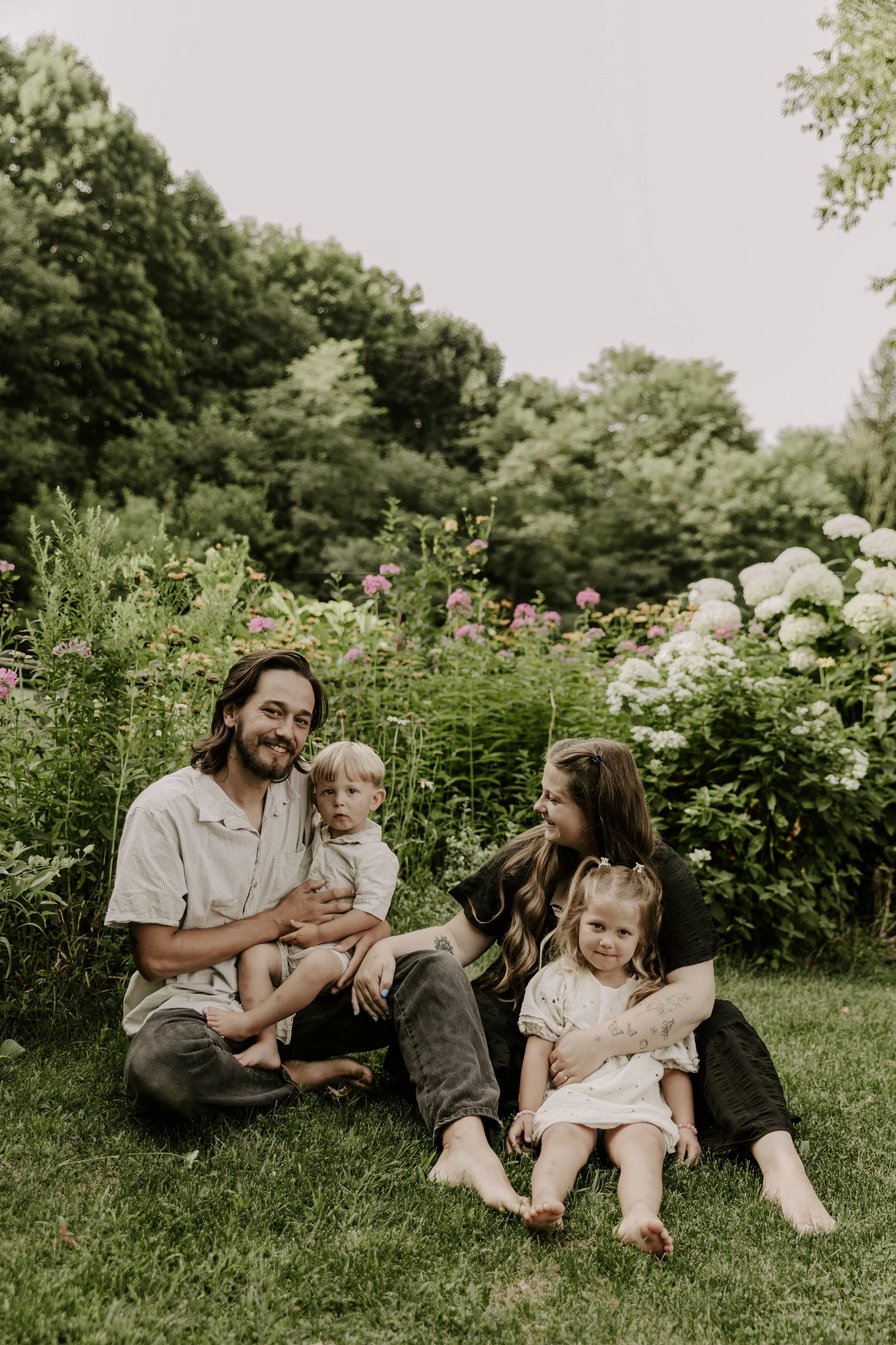 A family of five lying on the grass in a garden with flowering bushes and trees in the background. The man is holding a young boy, with a woman and a girl sitting nearby during maternity family photography package in Ottawa Valley Ontario.