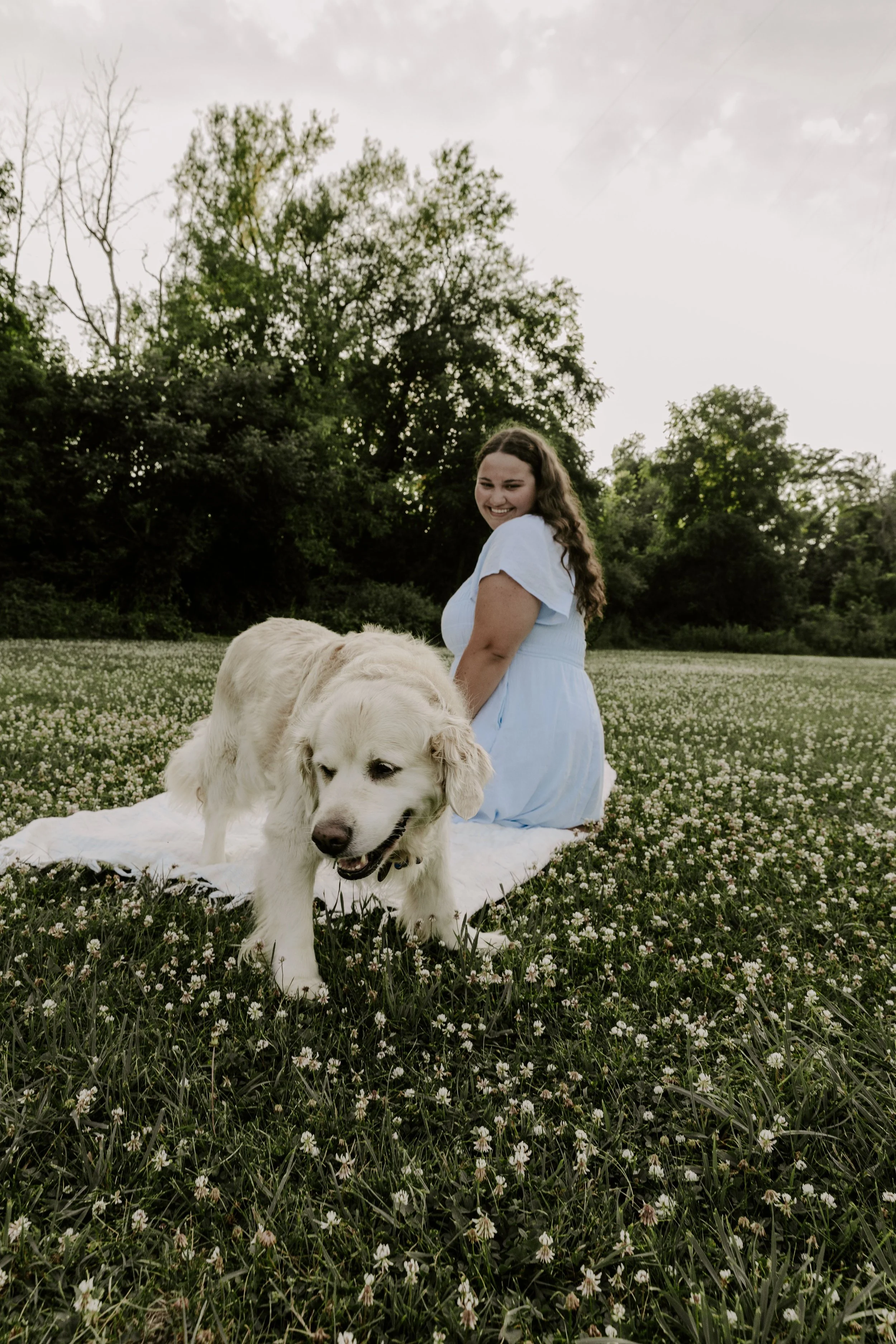 A woman photographer in a light blue dress sitting on a blanket in a field of small white flowers, with a golden retriever dog in the foreground. The background features trees and a cloudy sky.