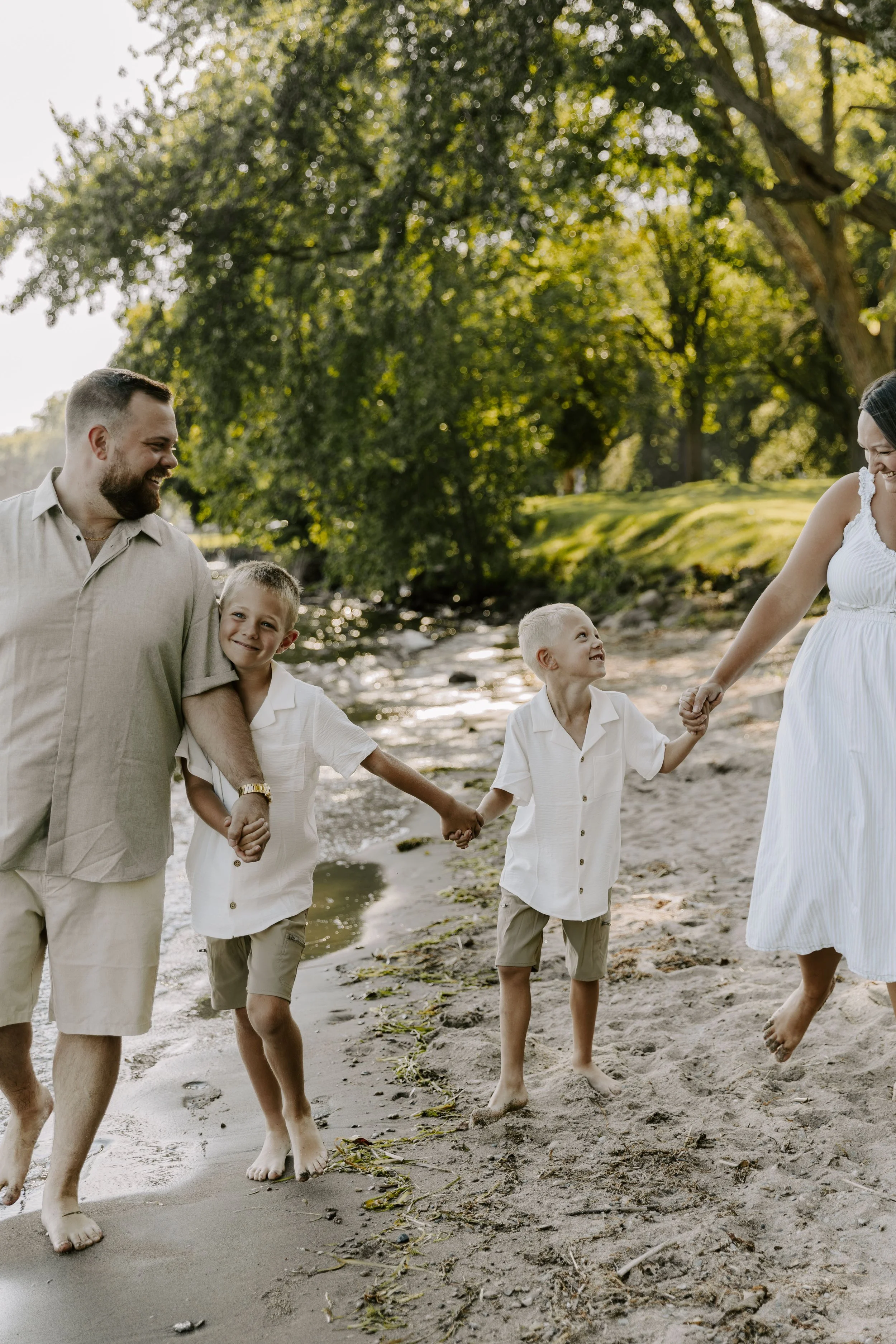 A family of five, including two adults and three children, walking hand-in-hand along a beach shoreline during daytime with trees and a small stream in the background for their family photography package in Ottawa Ontario.
