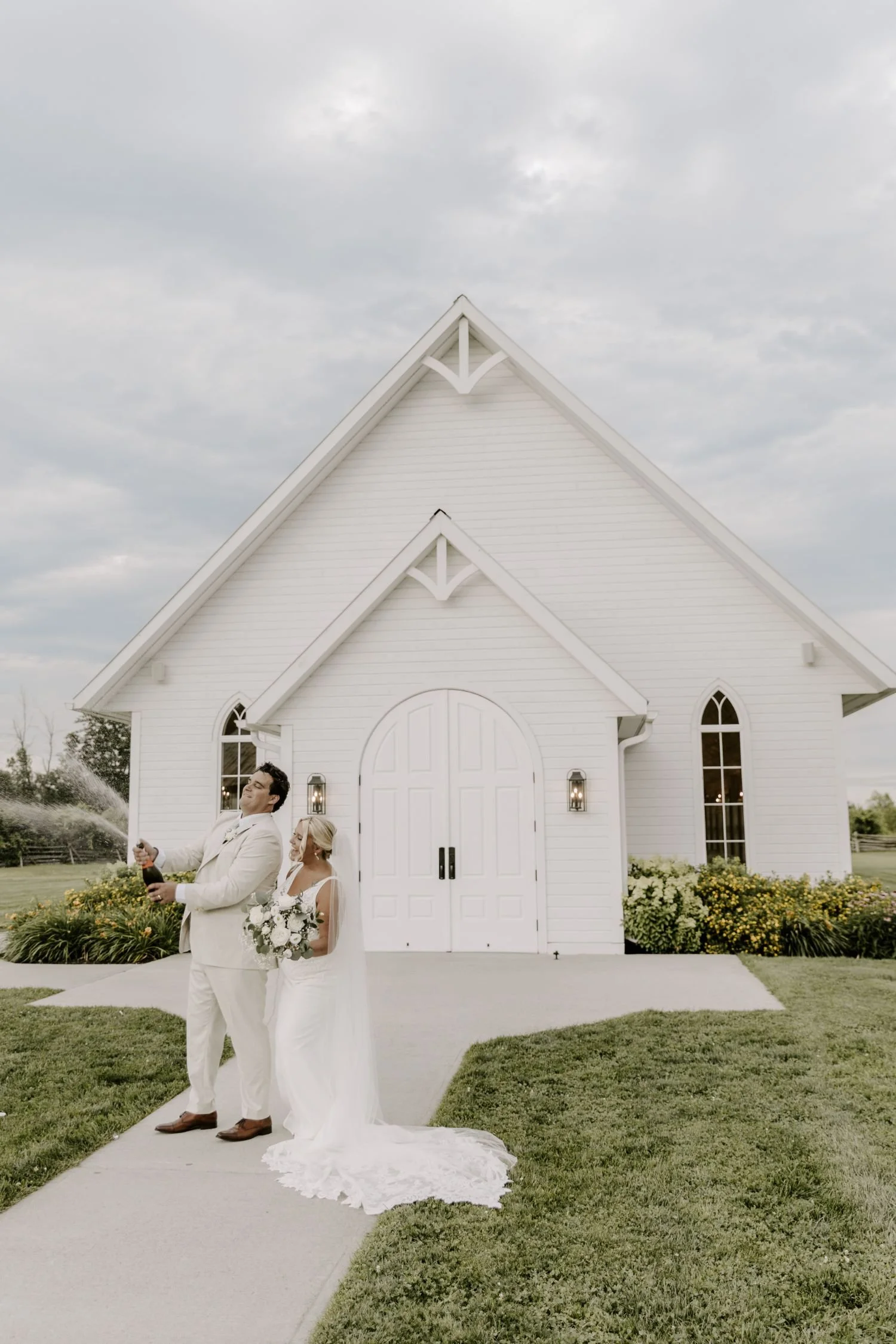 A bride and groom standing outside a white church with a pointed roof, celebrating a wedding. The groom is spraying champagne while the bride stands beside him holding a bouquet. The church has arched windows and a white door, surrounded by green grass and flower beds.