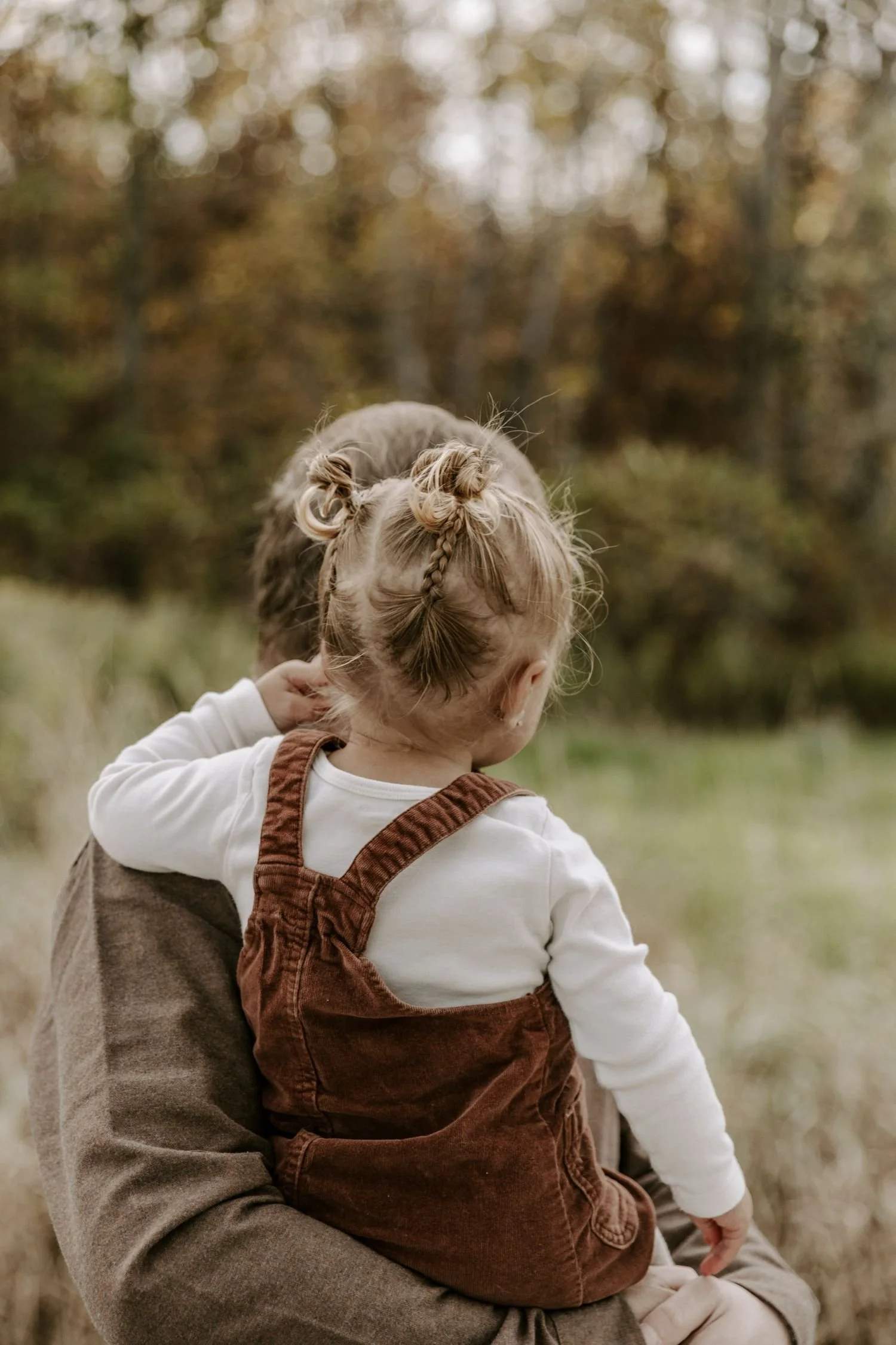 A young child with braided hair wearing brown overalls and a white long-sleeve shirt, sitting on someone's lap outdoors, with a blurred background of trees and autumn foliage for family photography package in Ottawa Ontario.