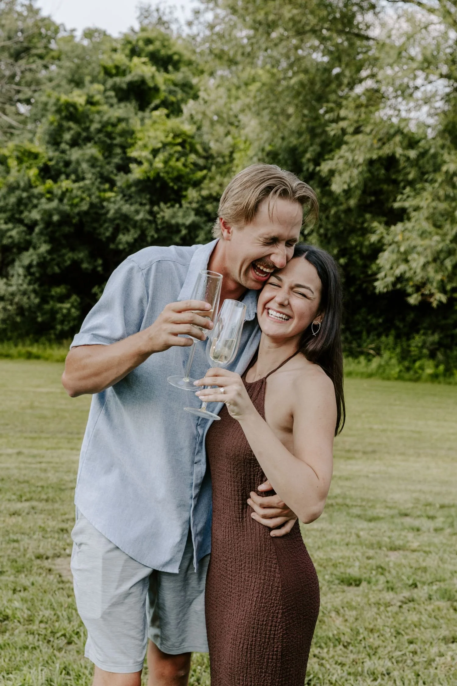 A man and woman are outdoors, smiling and holding champagne glasses, embracing each other during their engagement photography package in Brockville and Ottawa.