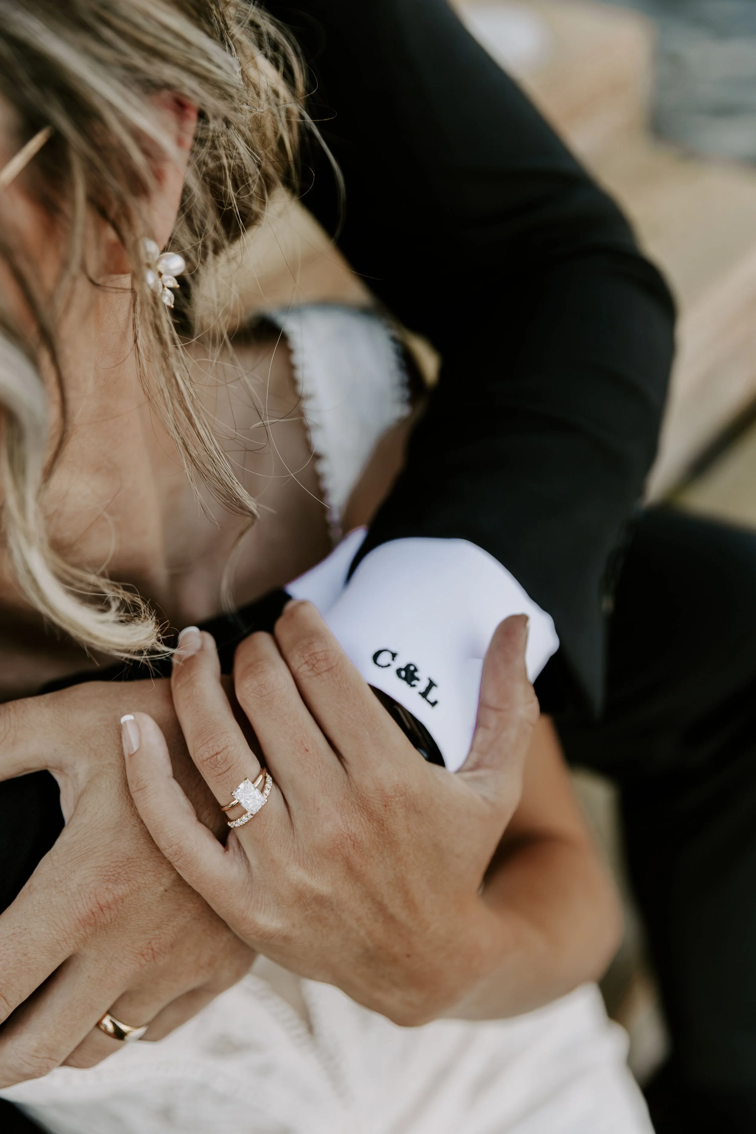 Close-up of a woman and a child hugging. The woman's hand, wearing a wedding and engagement ring, rests on the child's arm. The child is wearing a white top with black accents, and the woman has curly blonde hair and pearl earrings.
