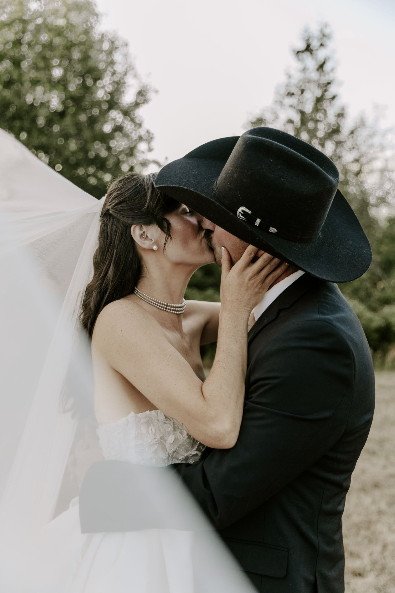 A bride and groom kissing outdoors, with the bride wearing a white gown and pearl necklace, and the groom in a black suit and large black hat, during daytime for their wedding photography package in Ontario